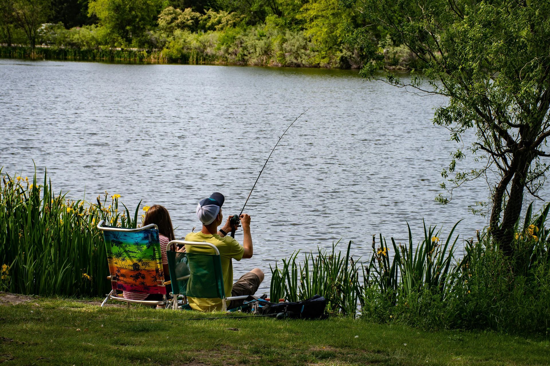 Couple fishing by a lake, seated in chairs, green grass and foliage in background.