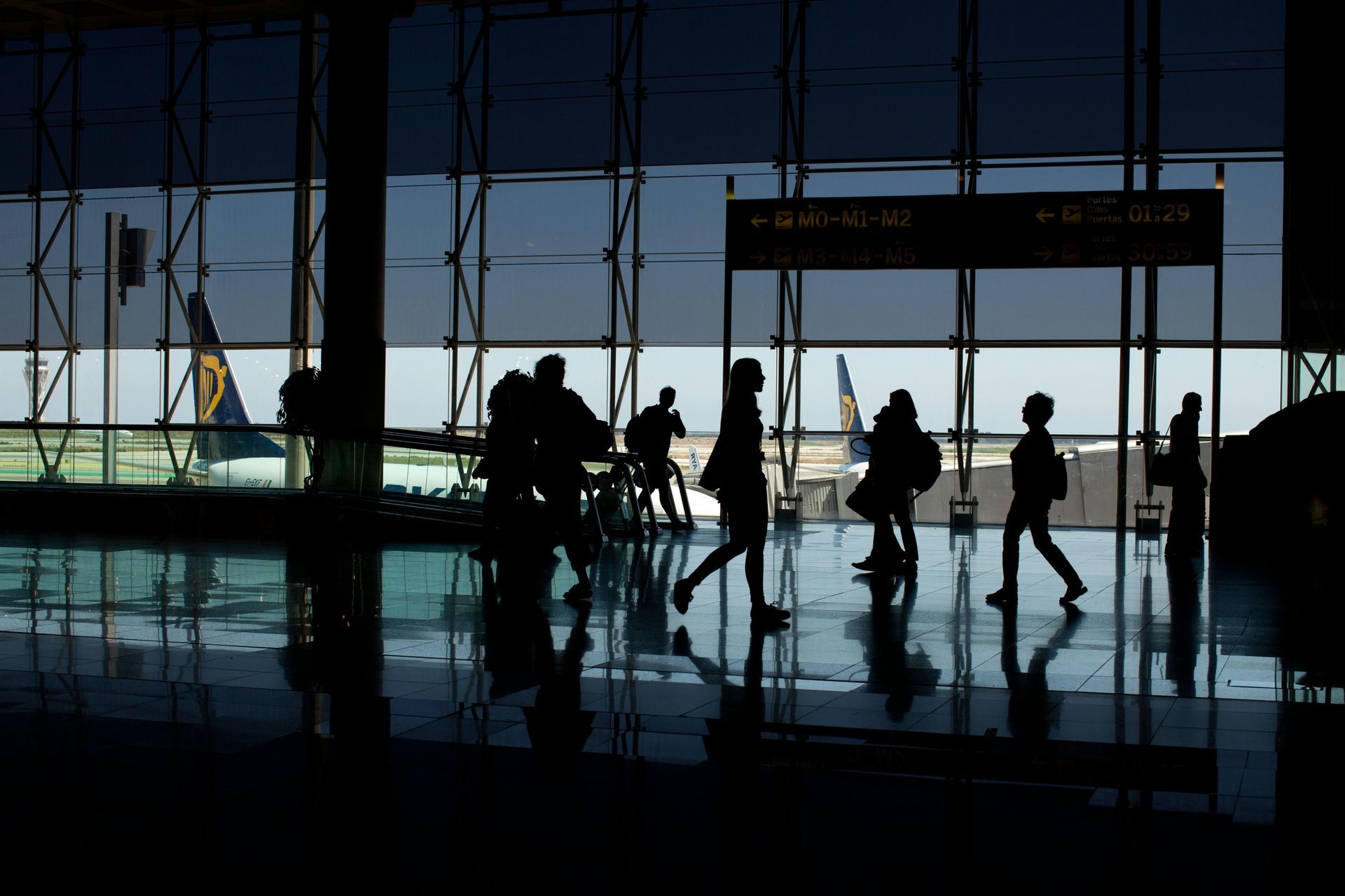 Silhouetted people walking in an airport terminal; a plane visible through windows.