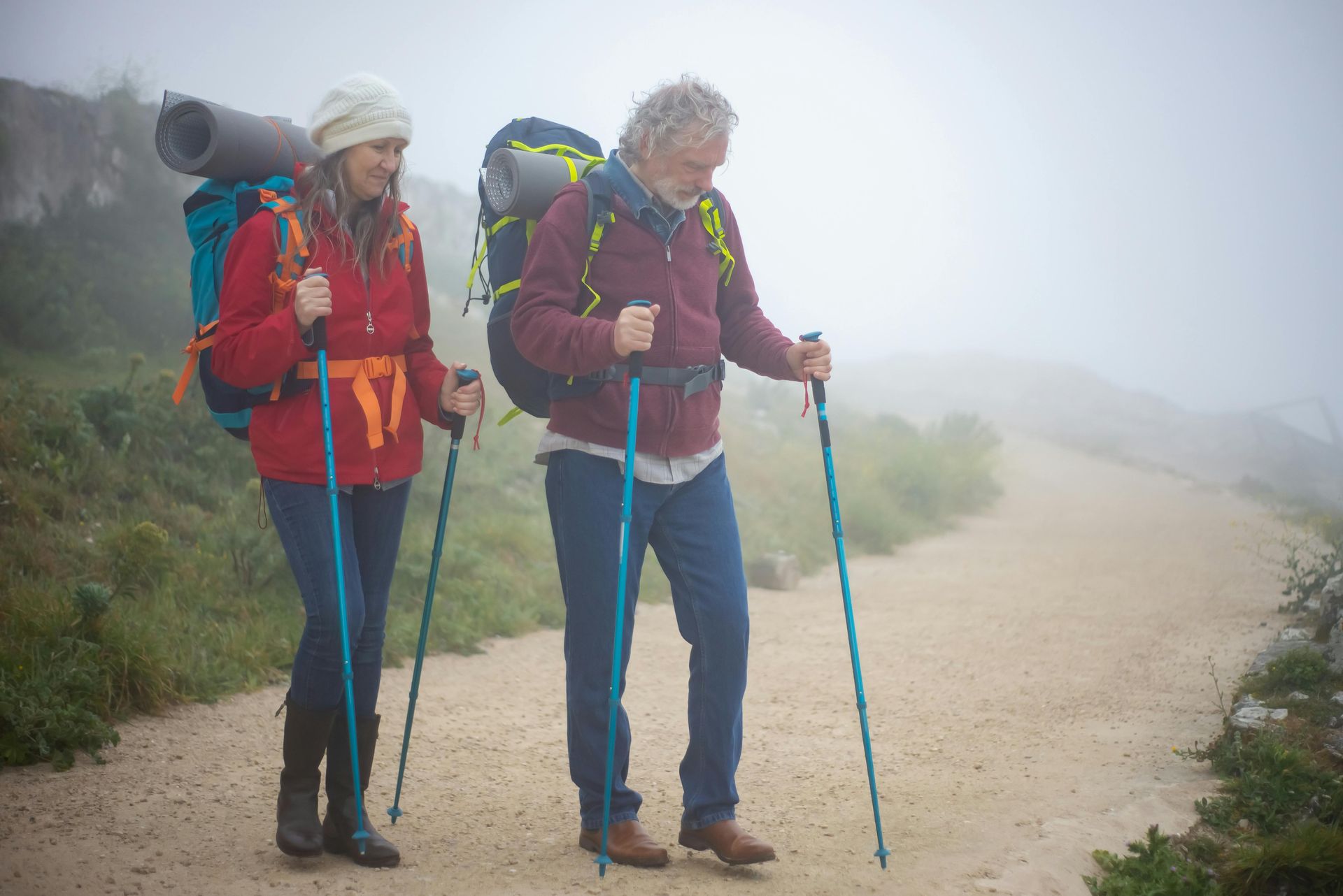 Two hikers, red jackets, backpacks, using trekking poles, walking path, foggy landscape.