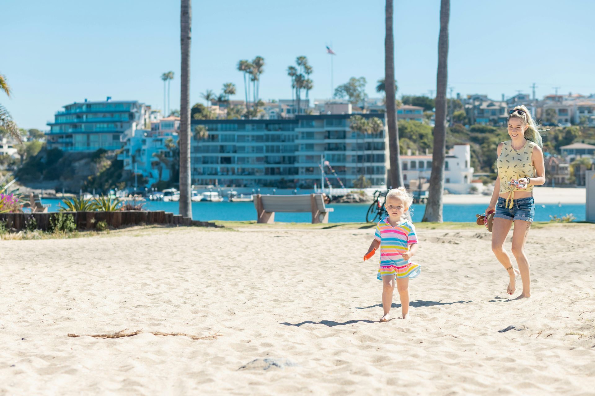 Mother and child at beach; child running towards camera, mother following. Buildings and water in background.
