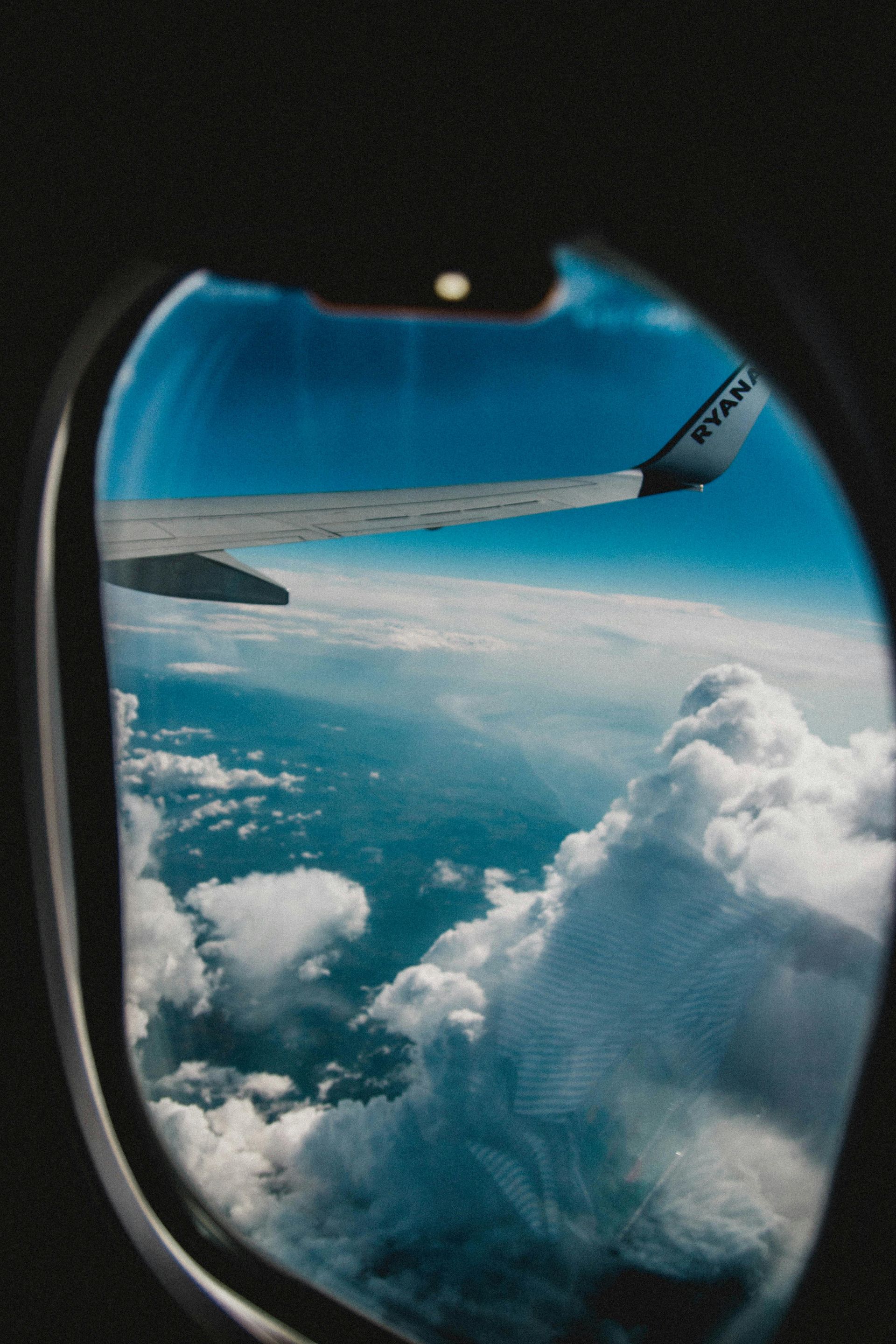 View from airplane window, wing in frame, blue sky, fluffy clouds below.