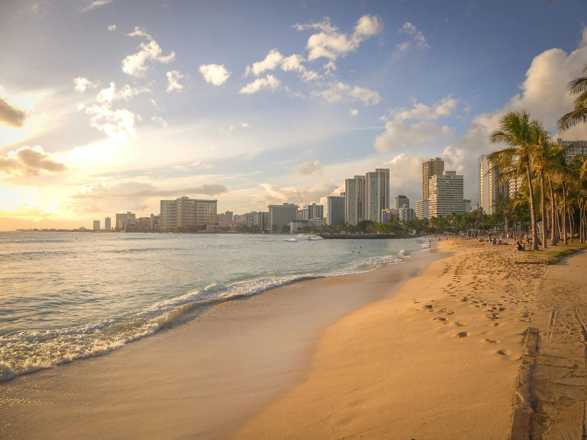 Beach scene with ocean waves, golden sand, city skyline, palm trees, and a bright sky.