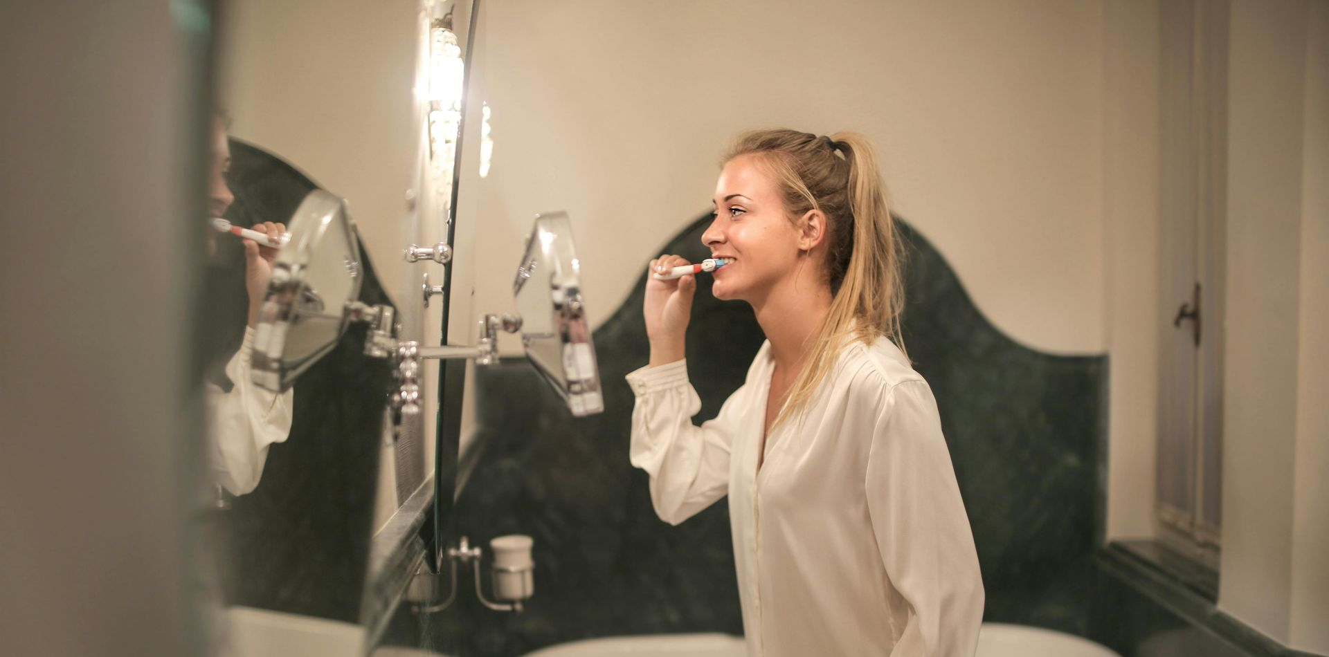 Woman in white shirt brushing teeth in bathroom, looking in mirror.