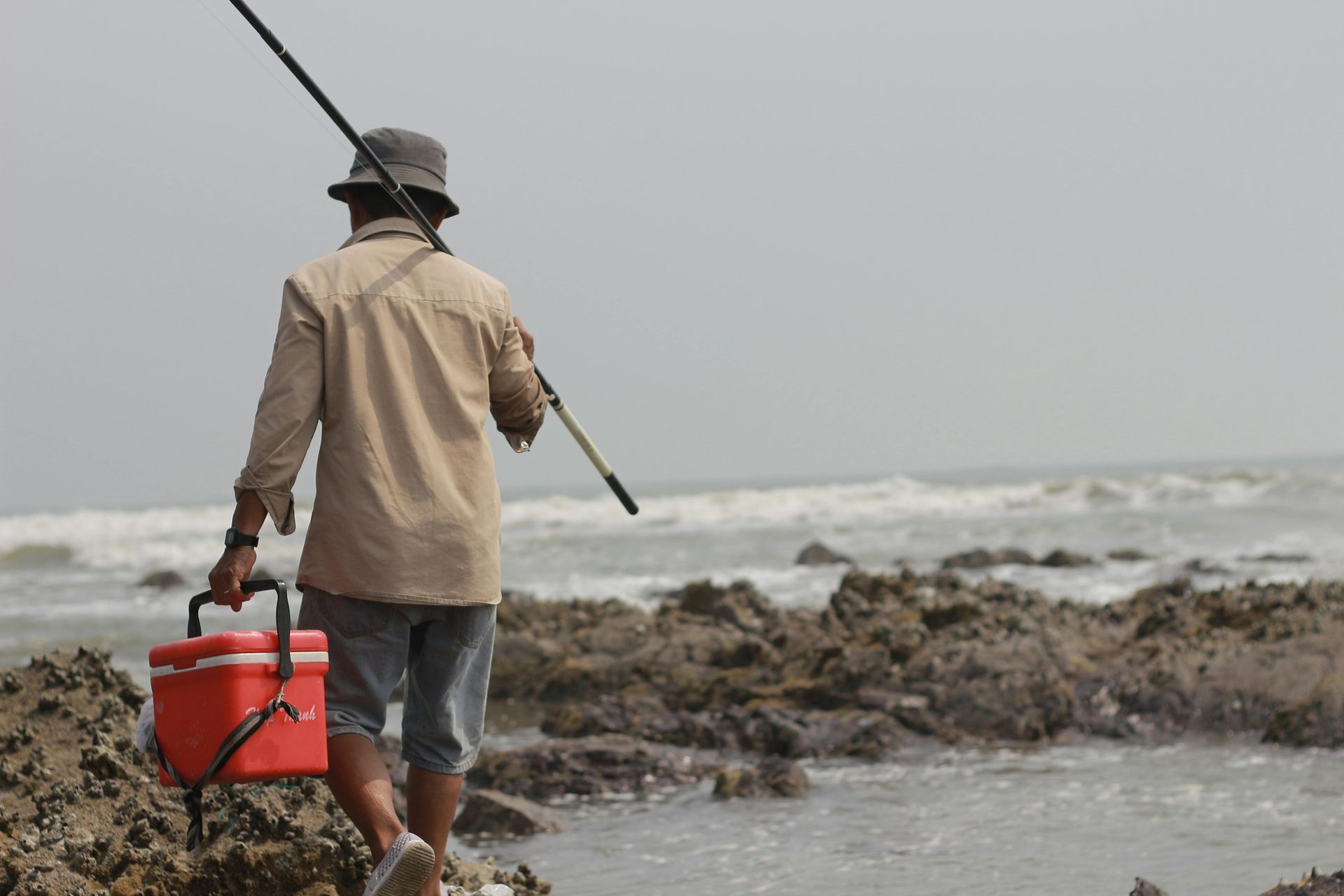 Fisherman carrying a red cooler and fishing rod walks on rocks toward the ocean.