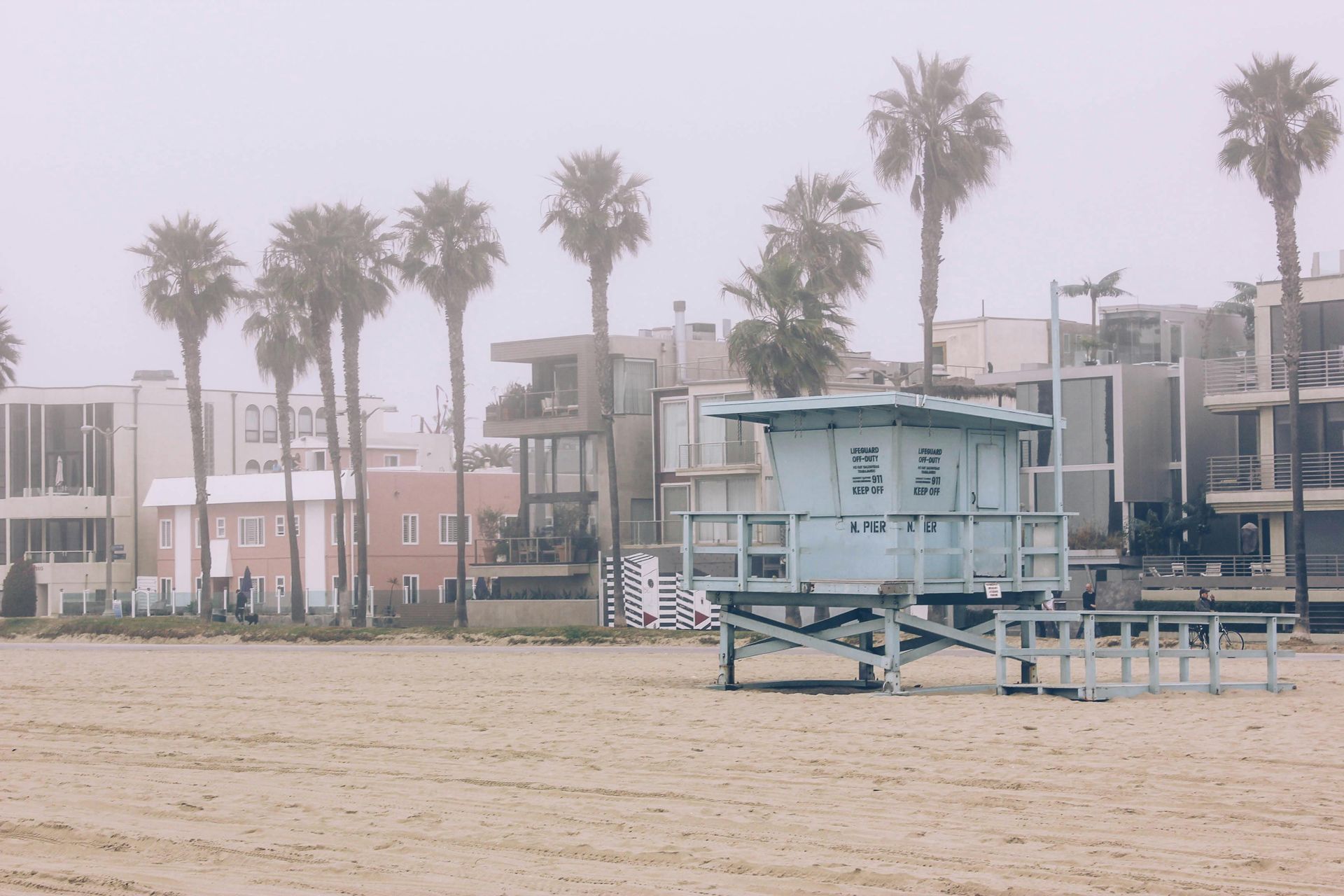 Beach with lifeguard stand, palm trees, and houses on a foggy day.