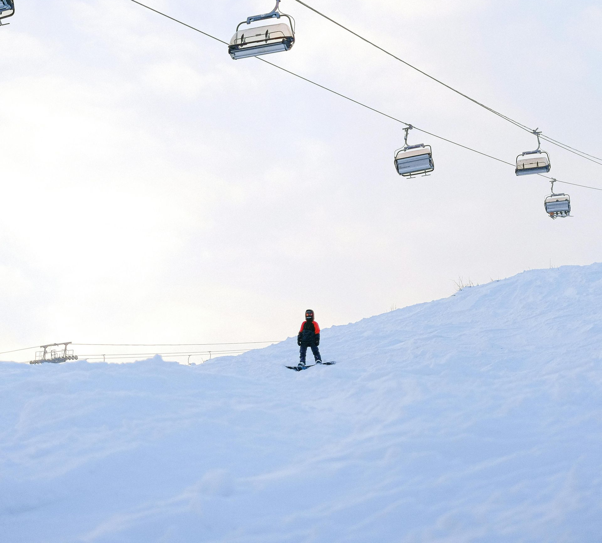 Skier on snow slope, under ski lift against overcast sky.