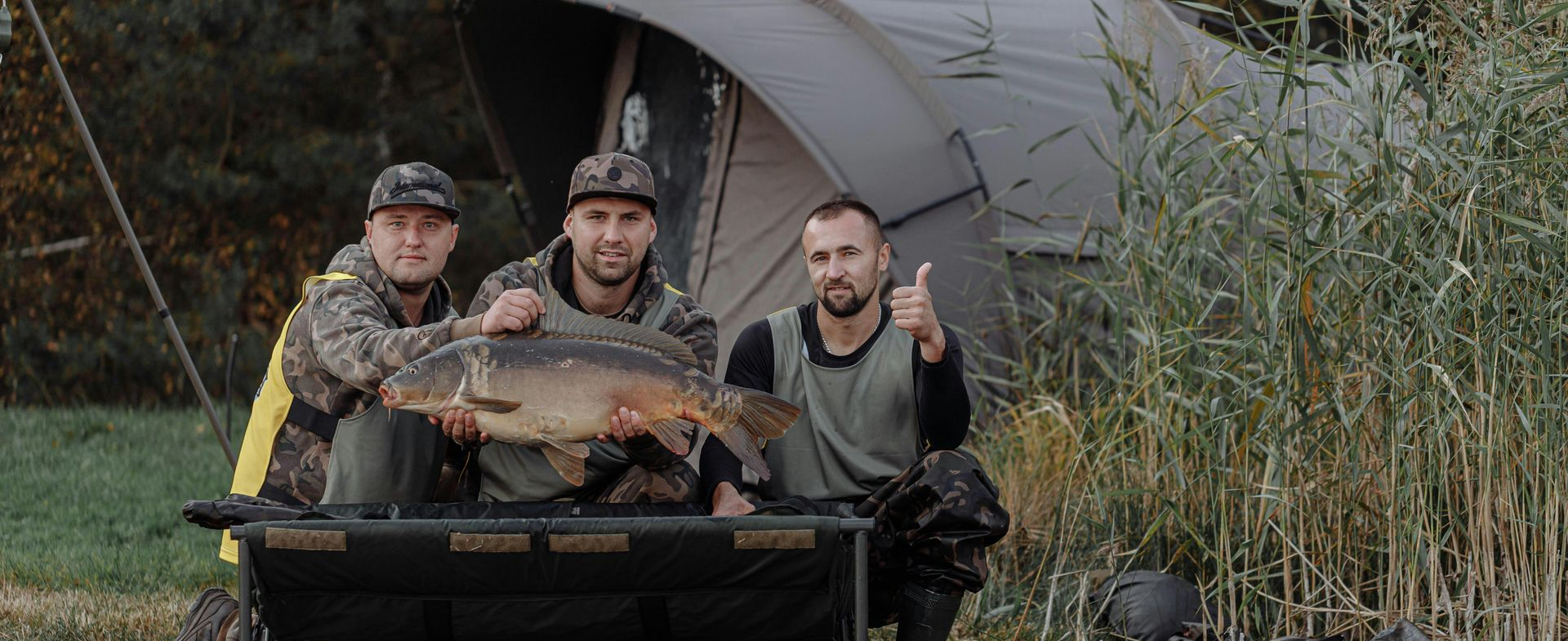 Three men holding a large carp fish by a lake, thumbs up, campsite in background.