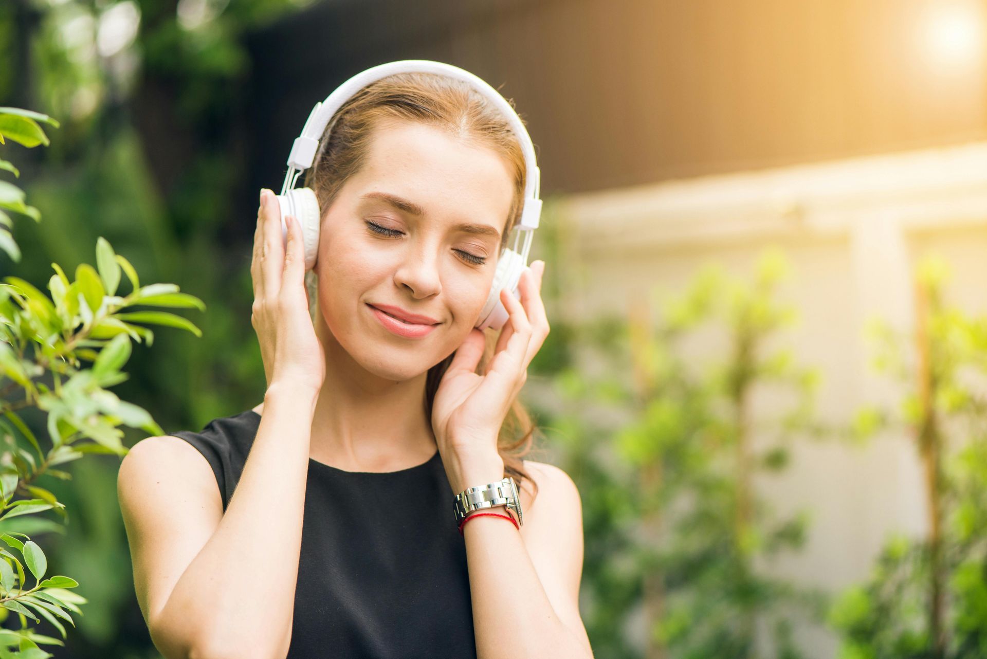 Woman with white headphones, eyes closed, smiling, enjoying music outdoors.