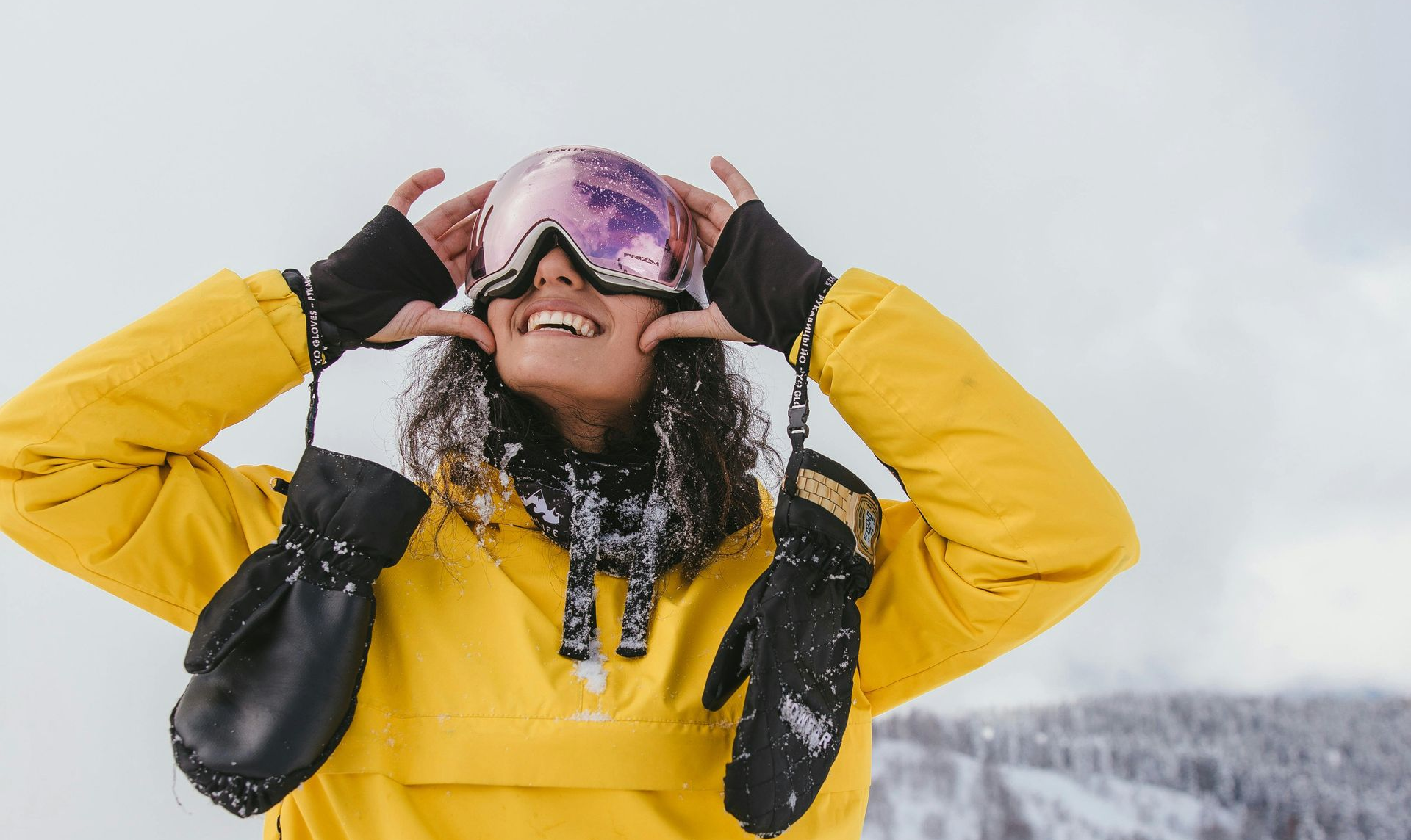 Person wearing ski goggles and yellow jacket smiles, touching face with mittened hands in snowy setting.