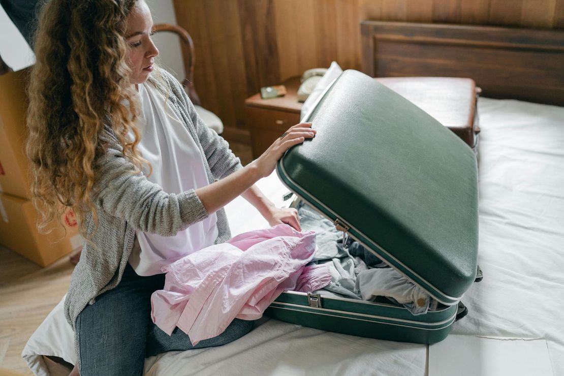 Woman packing a green suitcase on a bed with a pink shirt, in a room.