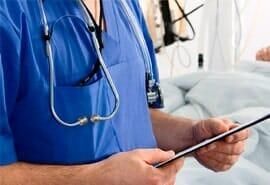 Un médico con un estetoscopio alrededor del cuello sostiene una tableta frente a un paciente en una cama de hospital.