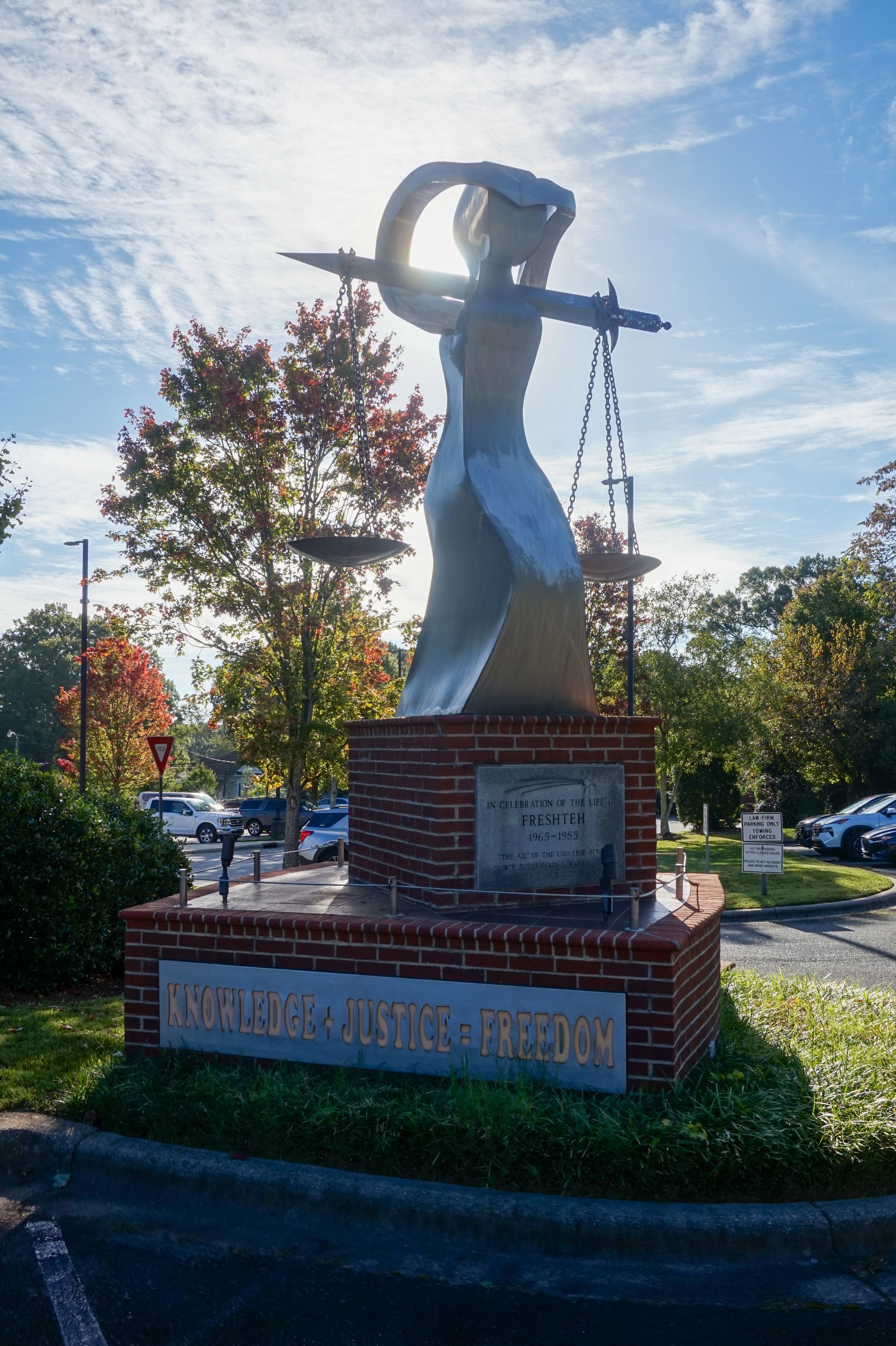 Statue of Lady Justice with scales, atop a brick base, in front of trees and a parking lot.