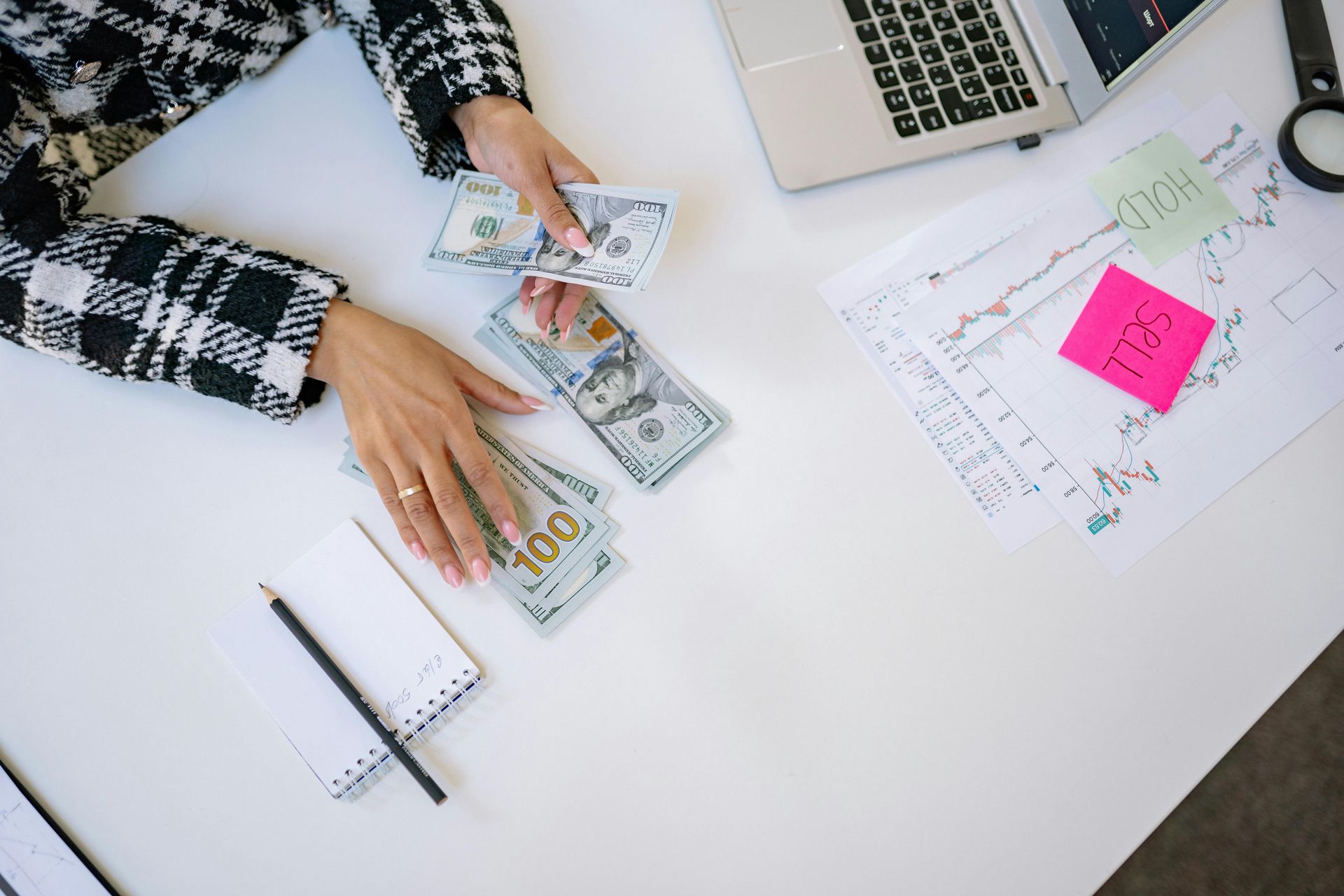 Hands counting cash at a desk with charts, notebook, and pink sticky note