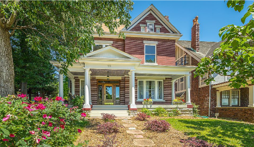 Victorian-style house with a front porch, pink flowers, and lush green trees
