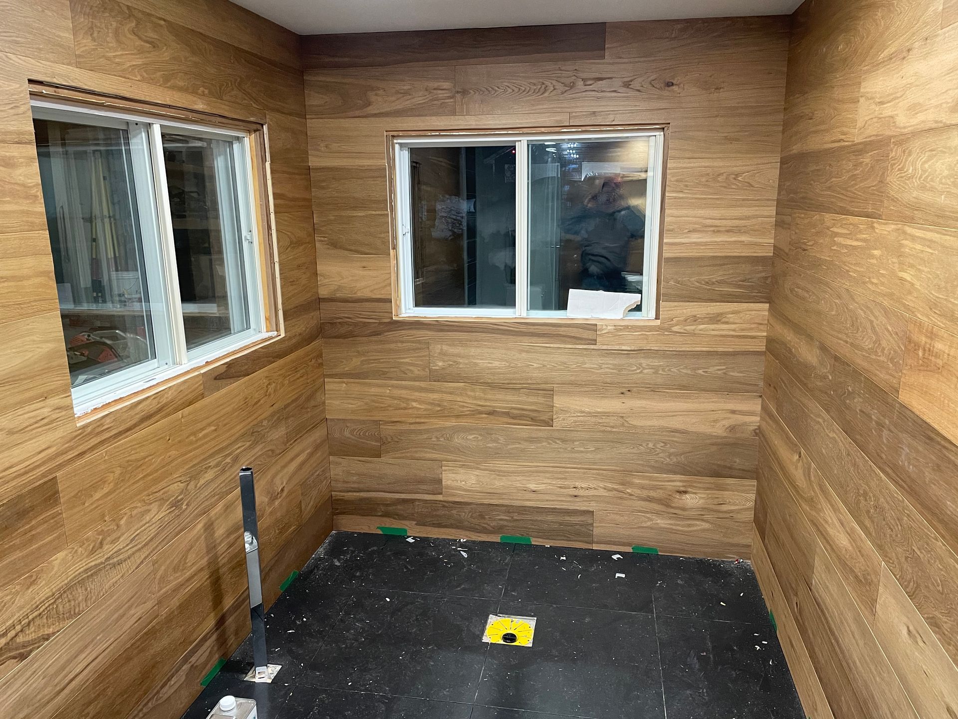 Pièce aux murs lambrissés et à deux fenêtres à cadre blanc ; sol sombre, non fini avec outils présents.