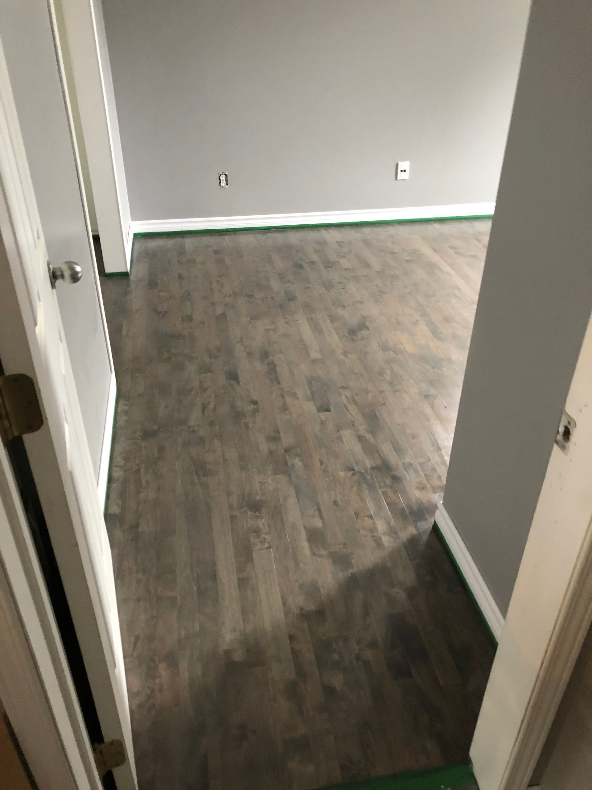 Vue d'une pièce avec parquet foncé, murs gris et moulures blanches ; du ruban adhésif vert souligne les plinthes.