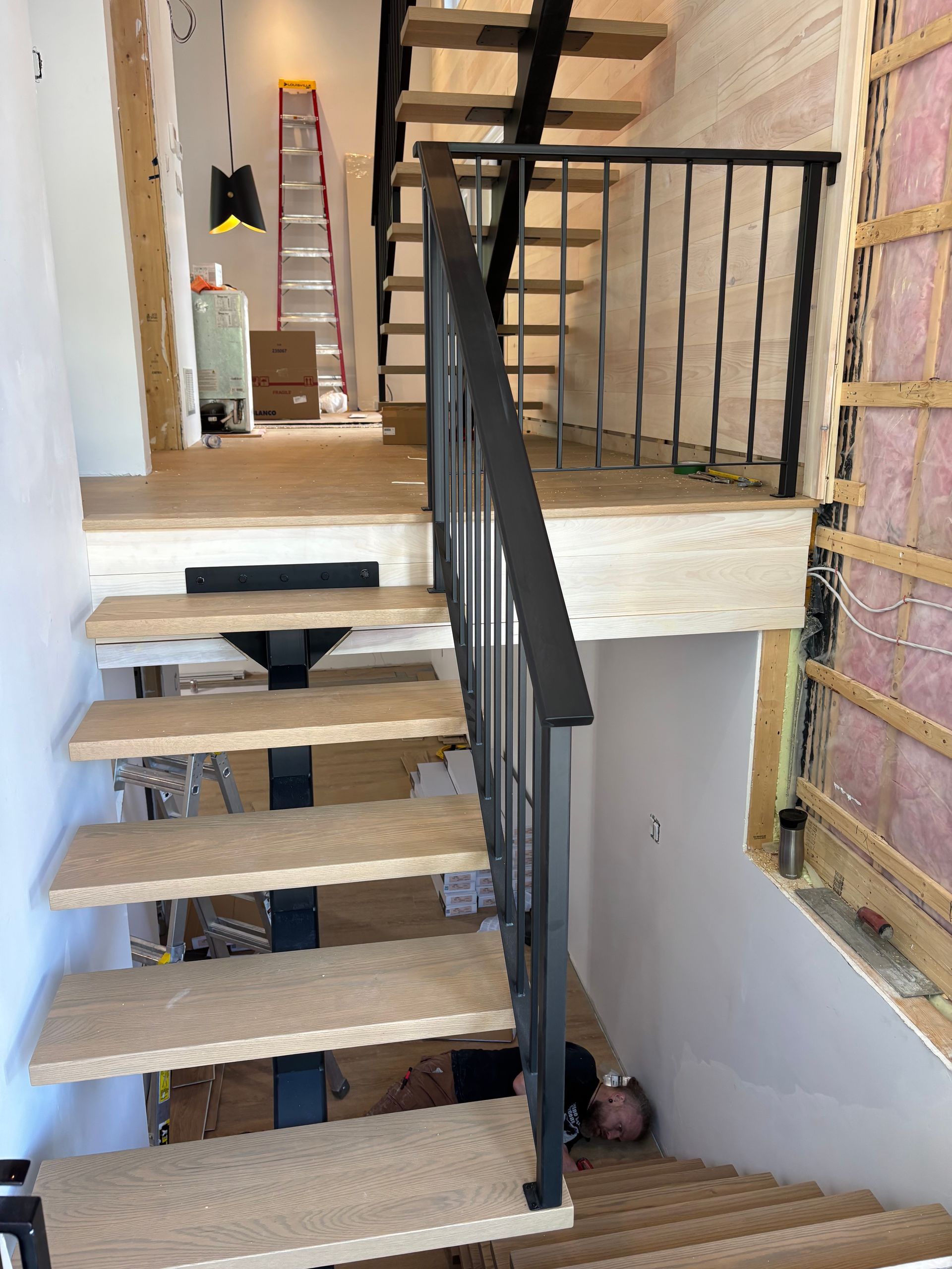 Escalier avec marches en bois clair, rampe en métal noir et matériaux de construction visibles.