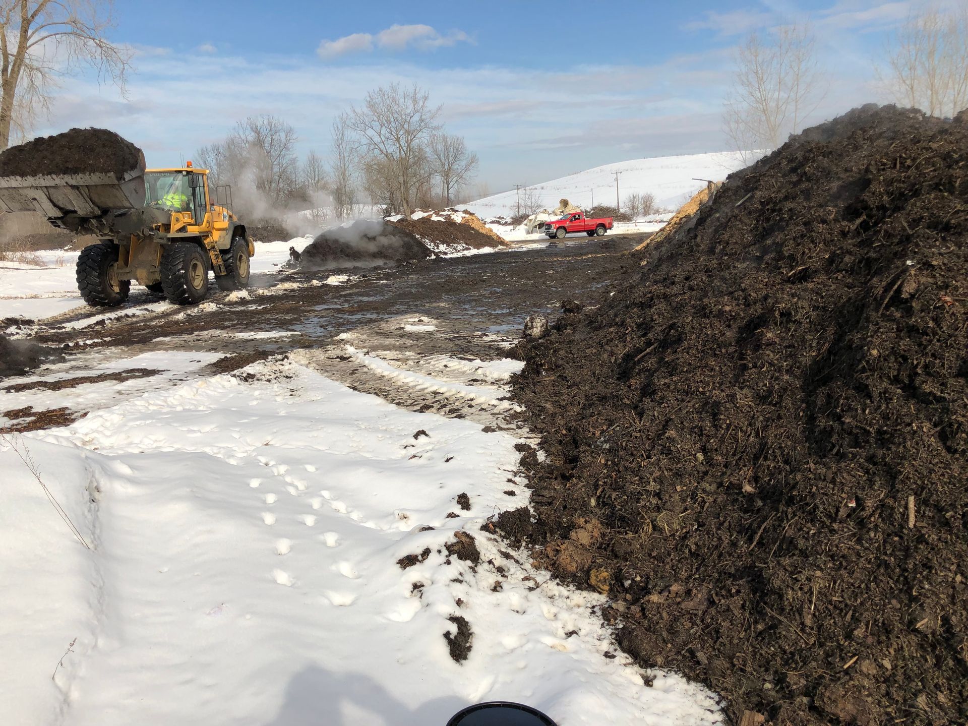 A front-end loader moves mulch on a snowy day, with large piles of mulch in a field.