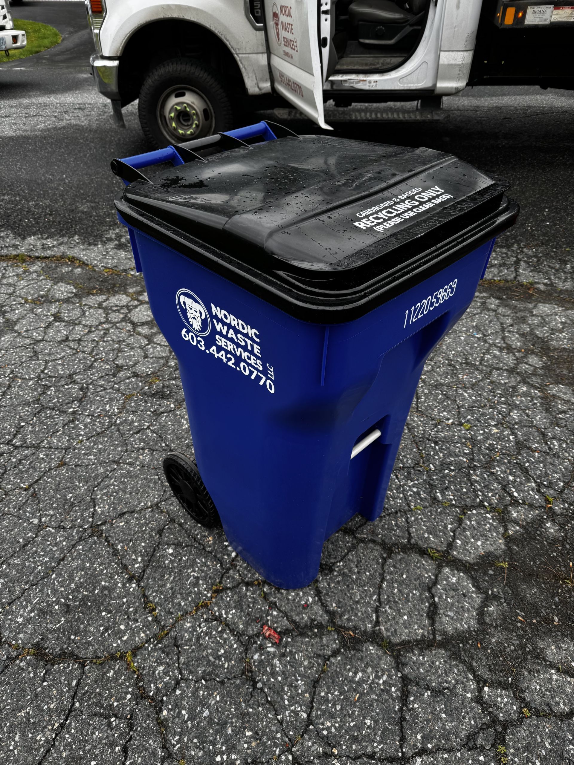 Blue trash can with black lid and wheels, in front of a white truck.