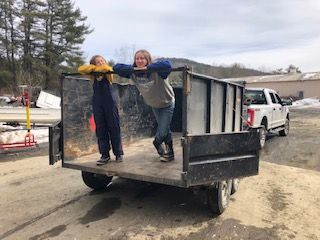 Two people pose in a trailer. Person on the left wears overalls and the other a sweatshirt. White truck in the background.