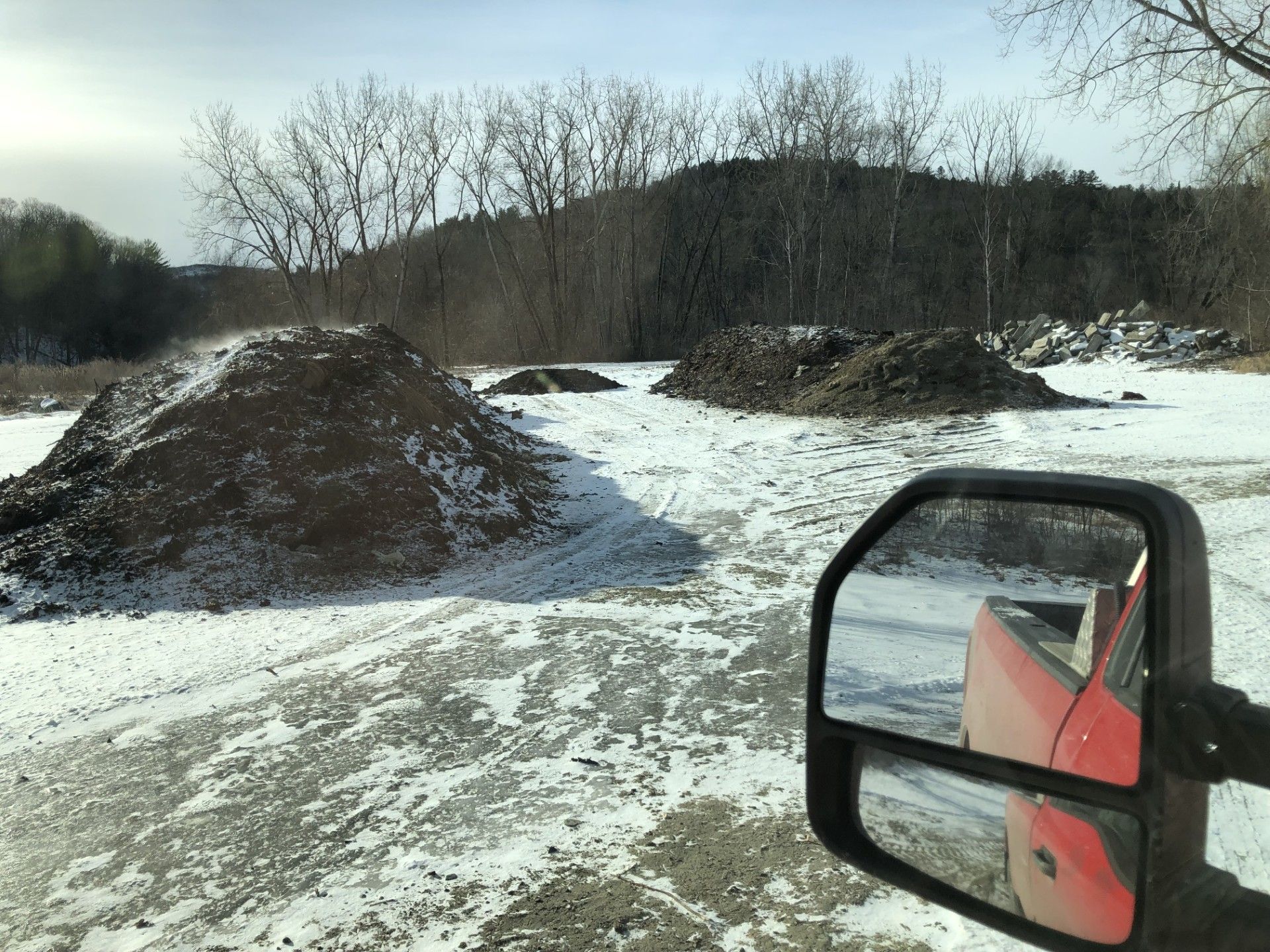 Snowy field with dirt piles; red truck mirror in foreground, trees and hill in background.