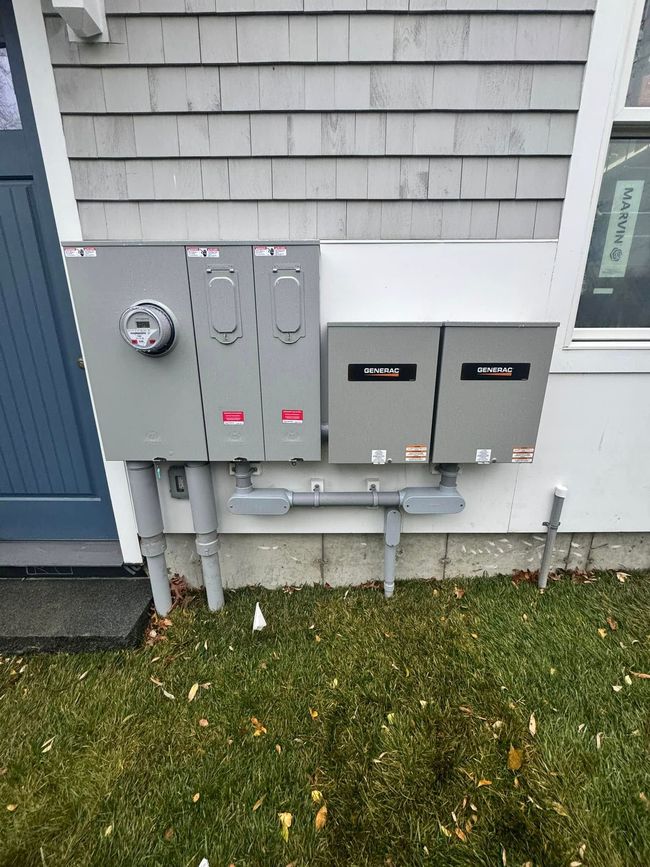 Electrical utility meters and gray service boxes mounted on the side of a house above grass.