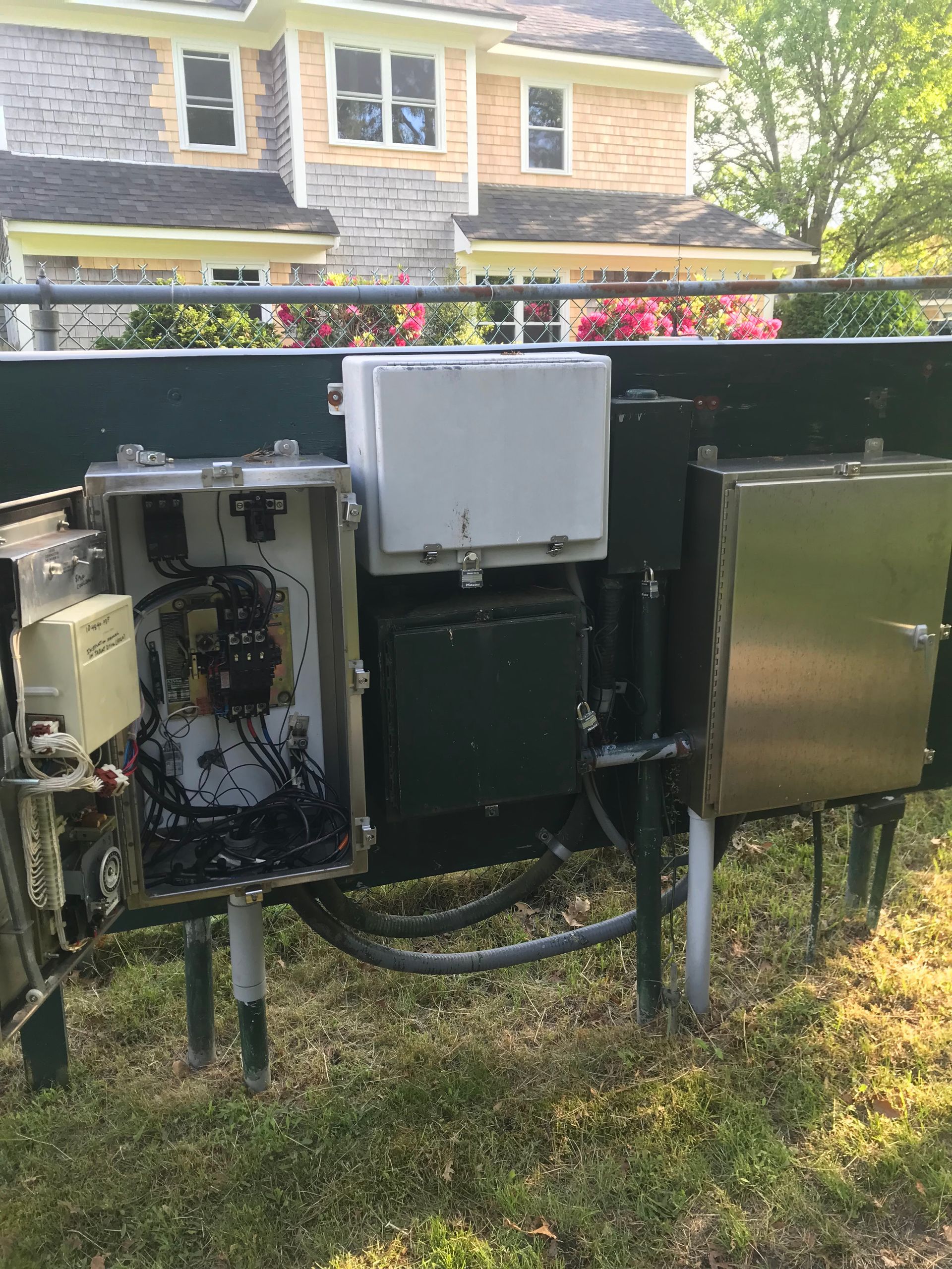 Open electrical utility boxes and meters mounted outdoors on a black fence, with a house and lawn behind them