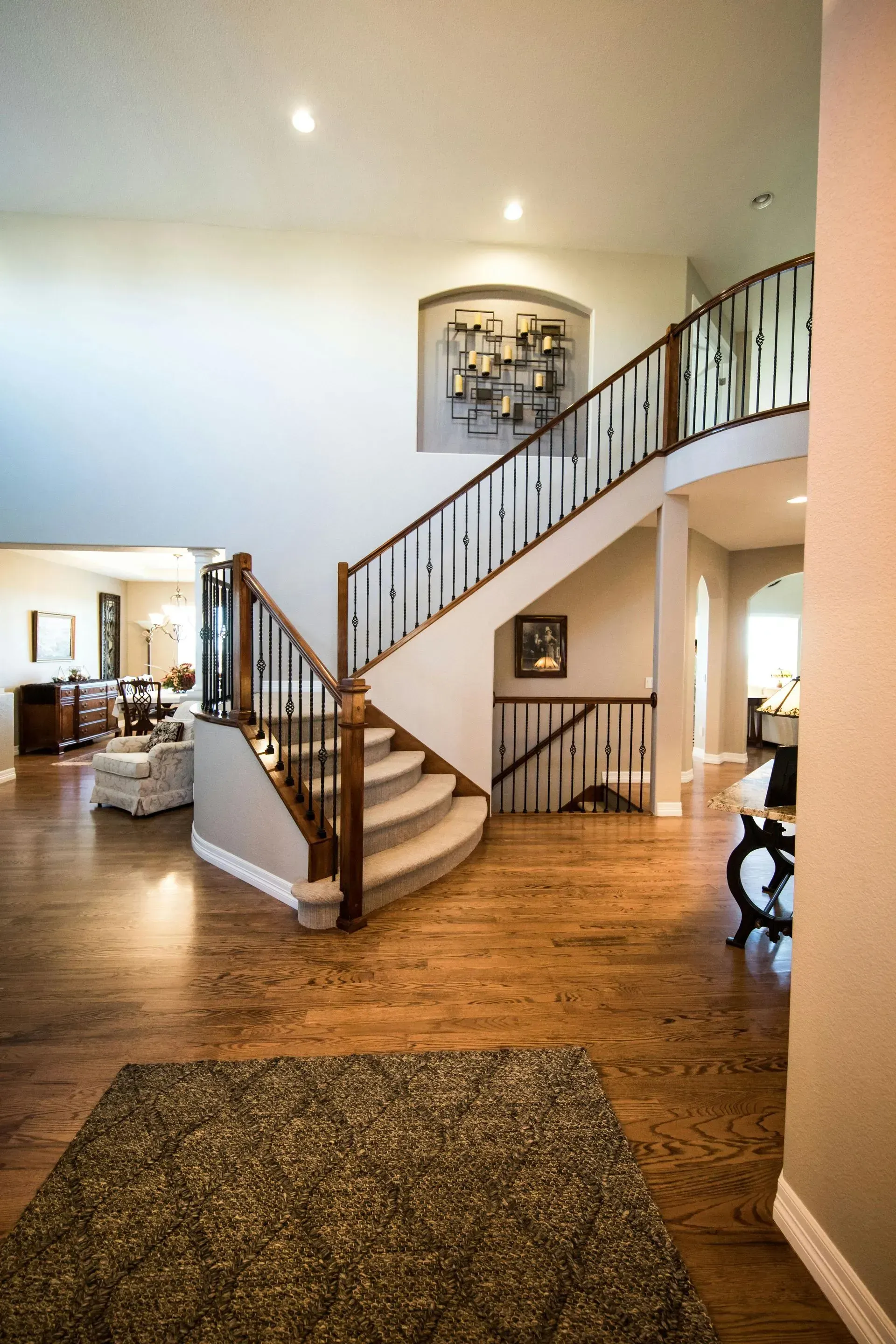 A grand foyer with hardwood floors, a carpeted curved staircase with dark railings, and an arched wall niche with art.