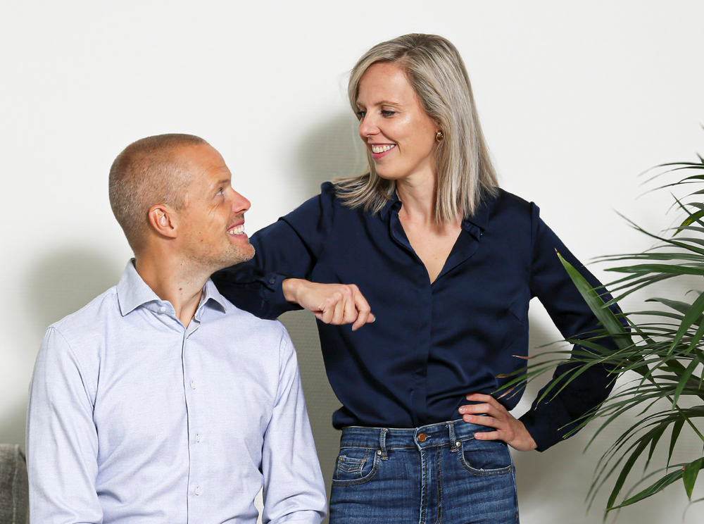 Man en vrouw lachen naar elkaar, vrouw leunt tegen de muur, binnenomgeving, neutrale achtergrond.