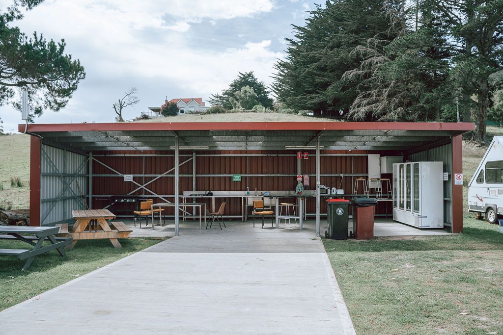 Covered outdoor kitchen with tables, grill, and fridges; red metal building with a green lawn.
