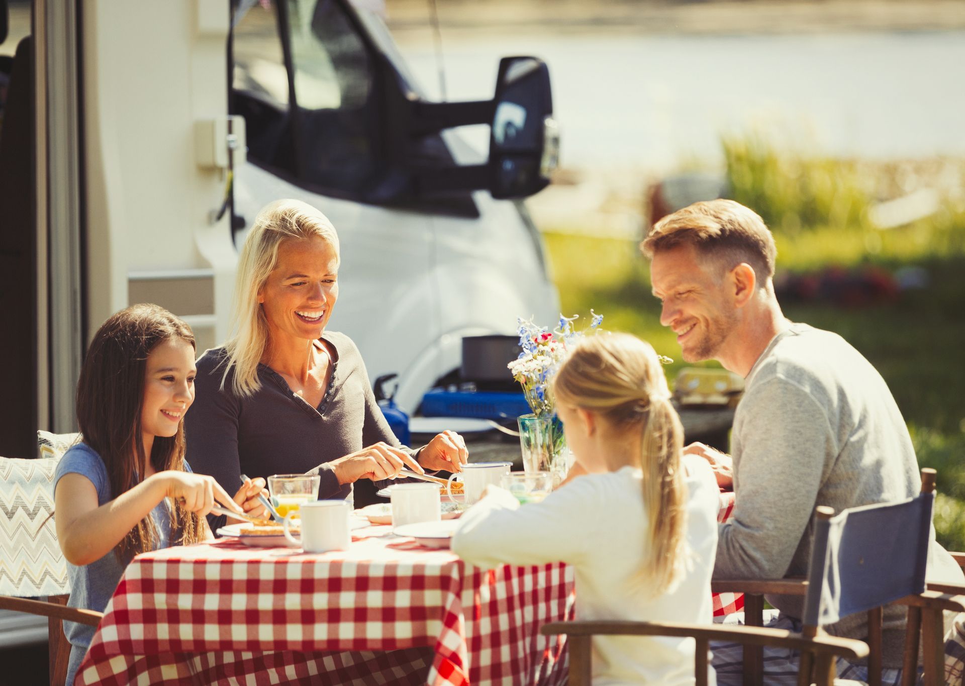 Family sitting at a picnic table outside a camper van enjoying drinks and snacks at a caravan park.