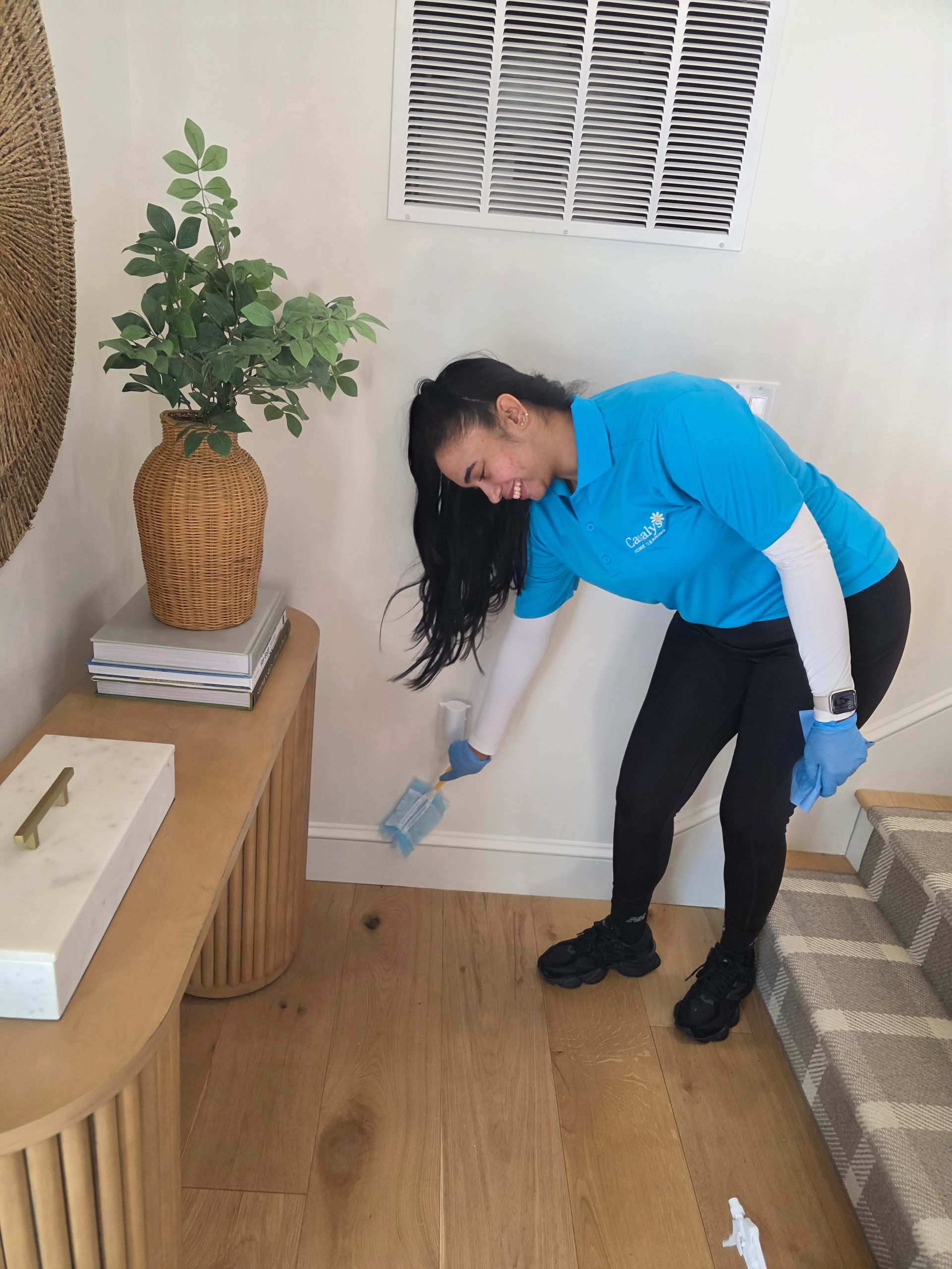 Cleaner in blue gloves dusting a wall beside a wicker plant and striped sofa in a living room