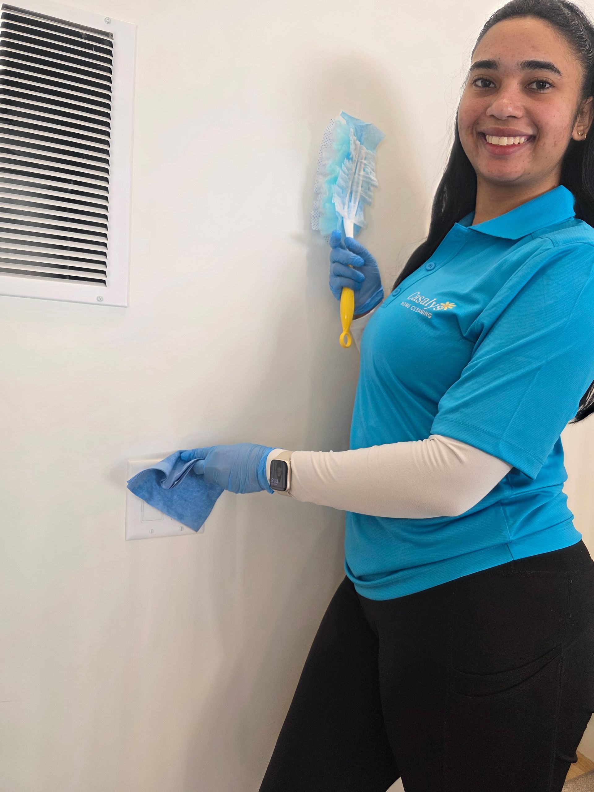 Woman in blue polo cleaning a white wall with a cloth and spray bottle, smiling.
