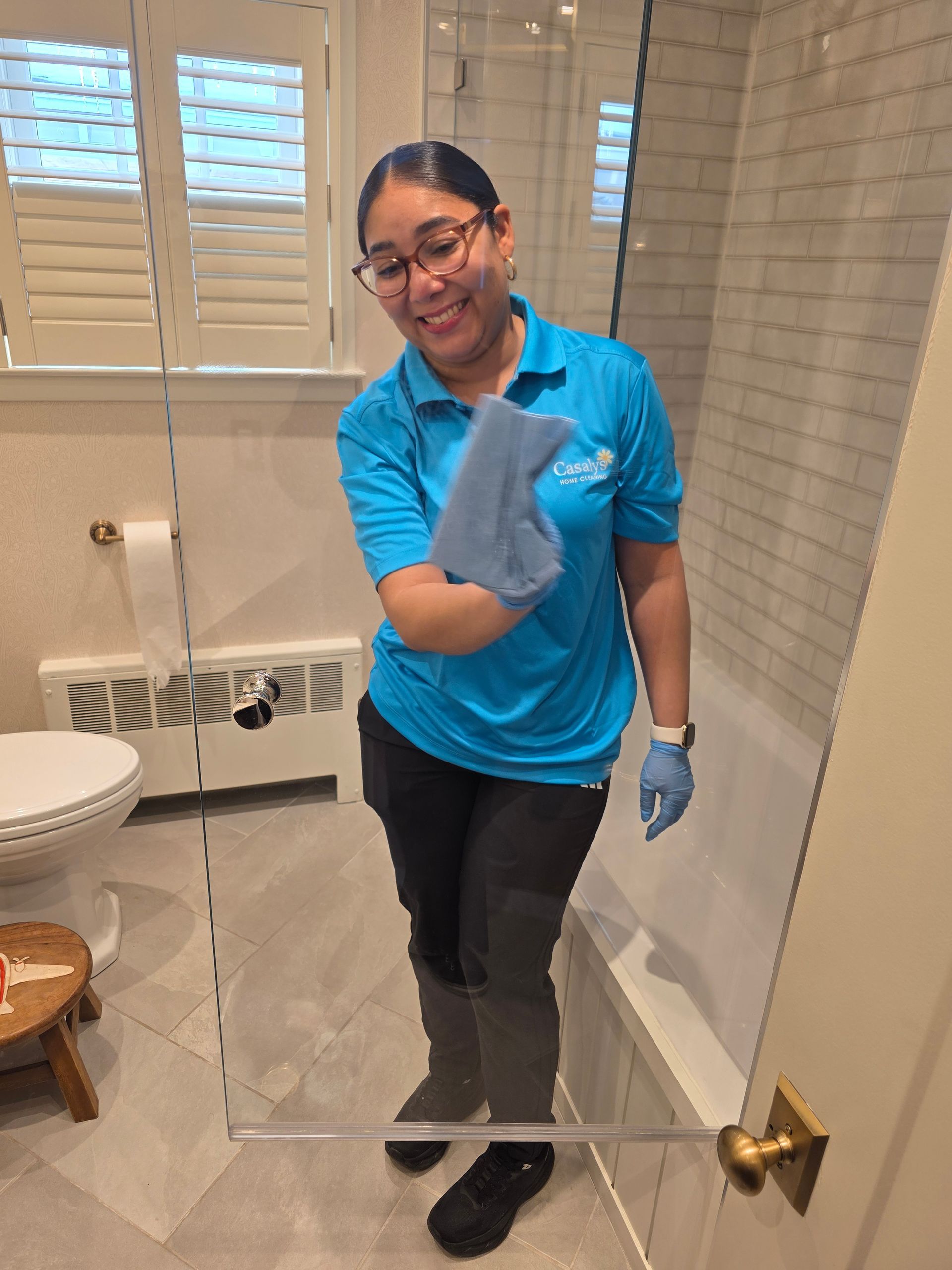 Person in a blue shirt and gloves cleaning a bathroom mirror in front of a sink and toilet.