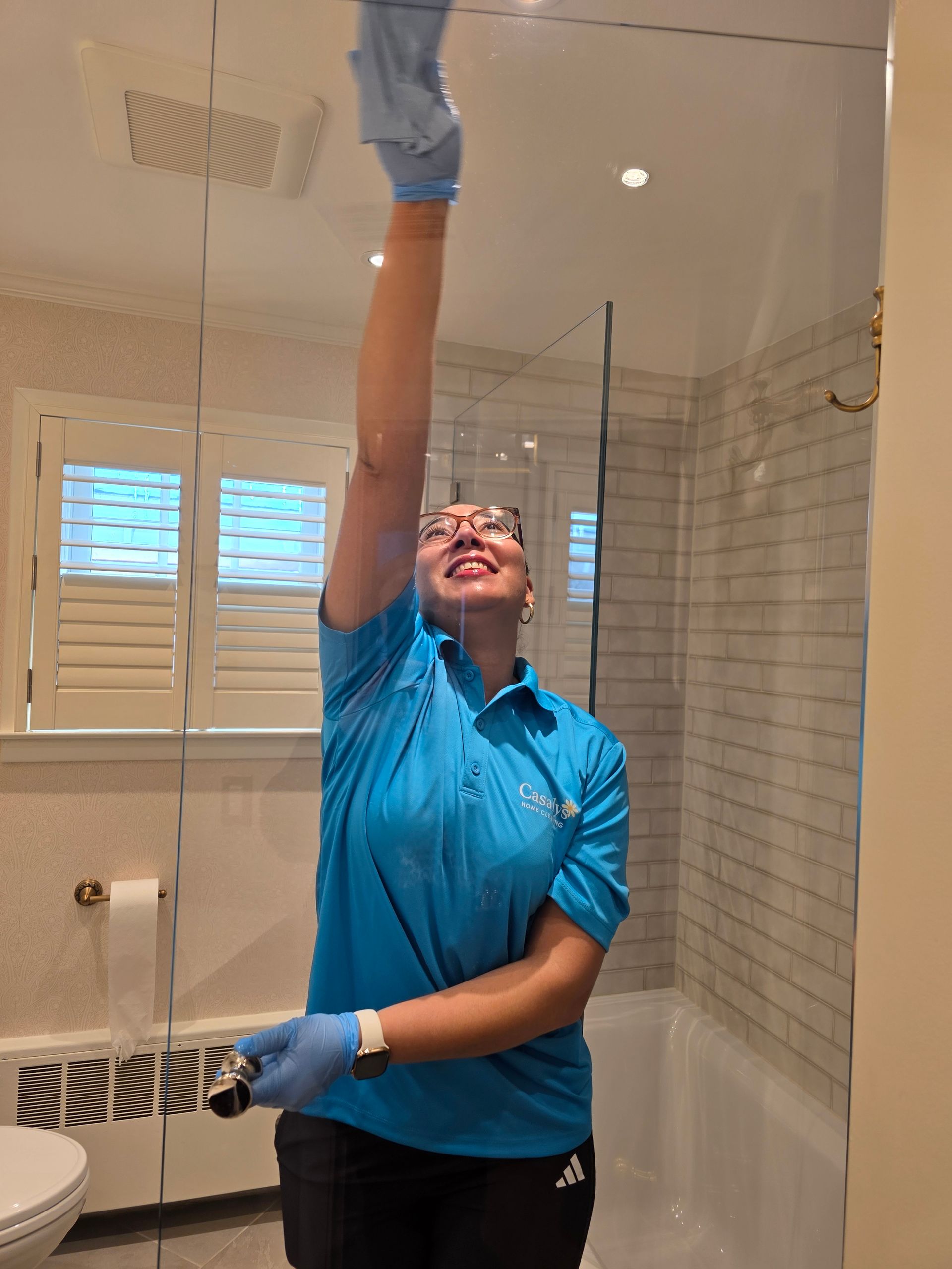 Person in blue shirt cleaning a shower glass wall with a squeegee, reaching overhead in a bathroom.