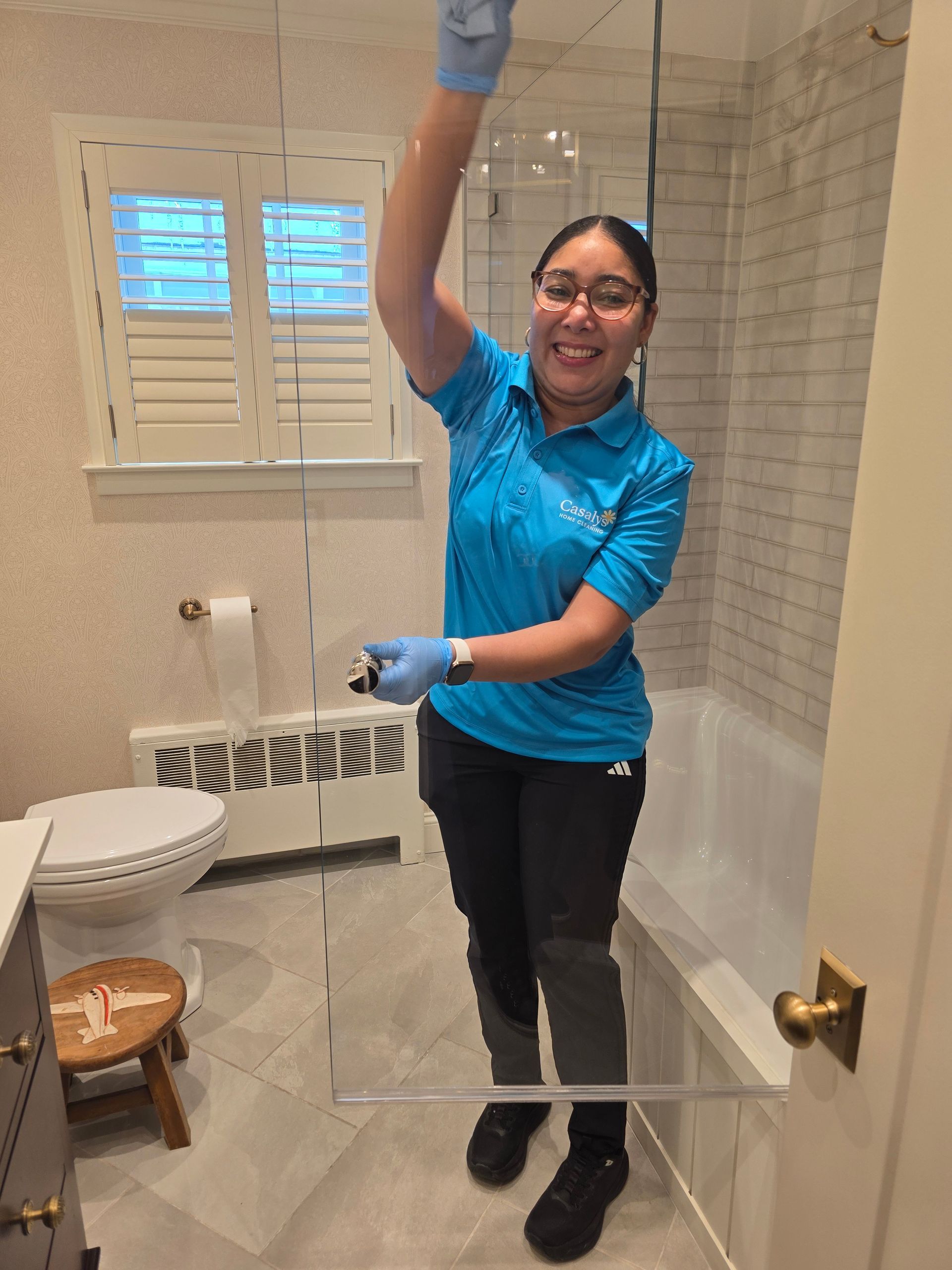 Person in blue shirt cleaning a bathroom shower, smiling and holding a spray bottle.