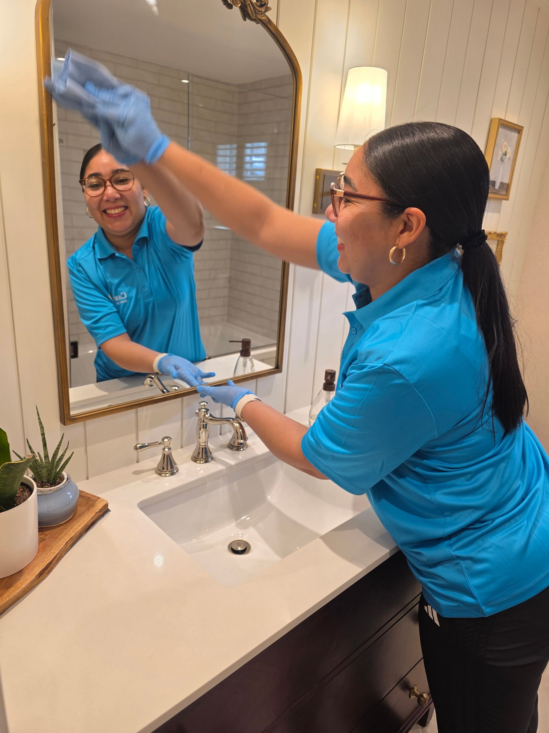 Person cleaning a bathroom mirror and sink with gloves, wearing a blue shirt.