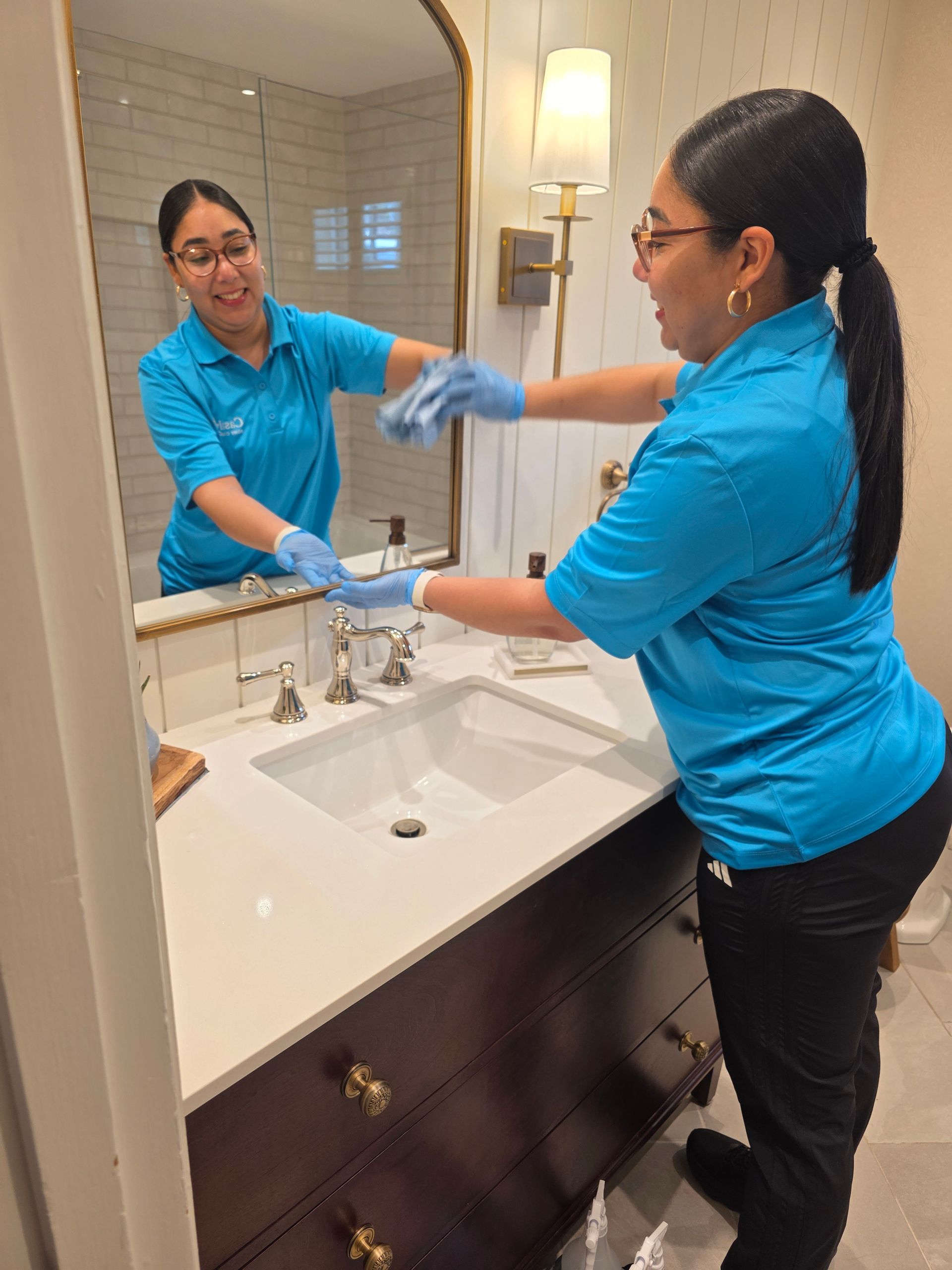 Woman cleaning a bathroom sink and mirror, wearing blue gloves and a blue shirt.