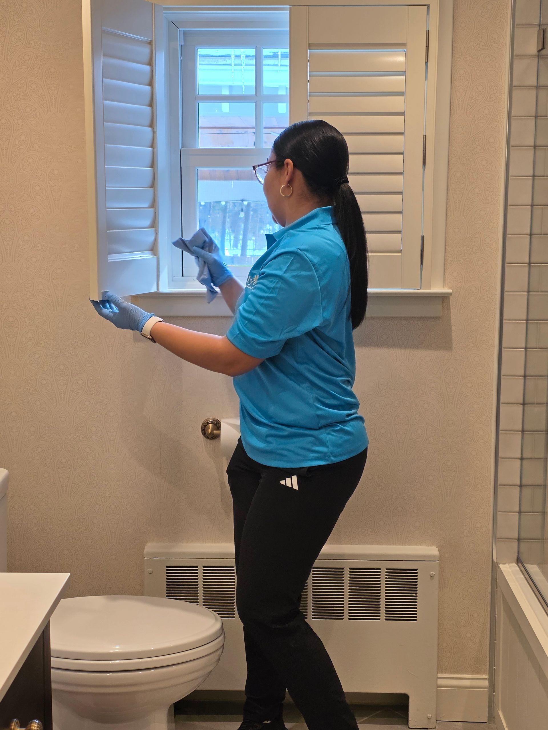 Person cleaning a bathroom window with blue gloves, standing by a toilet and vent.