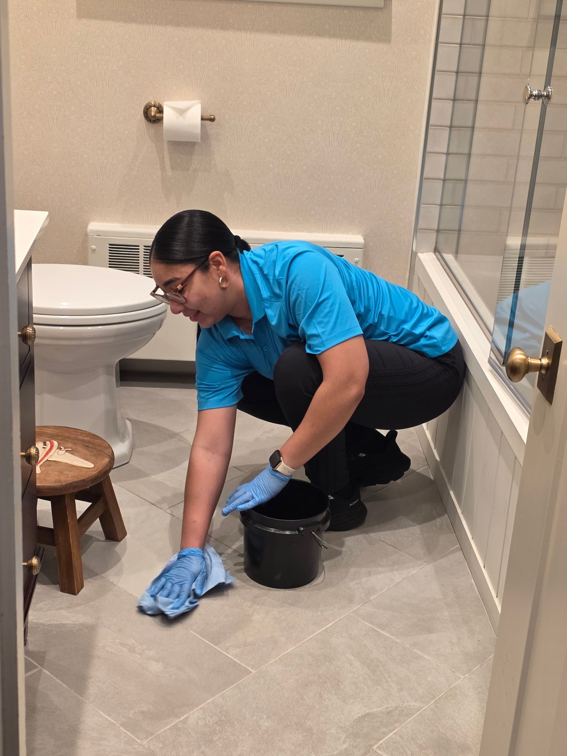 Person cleaning a bathroom floor with a blue cloth and bucket beside the toilet.
