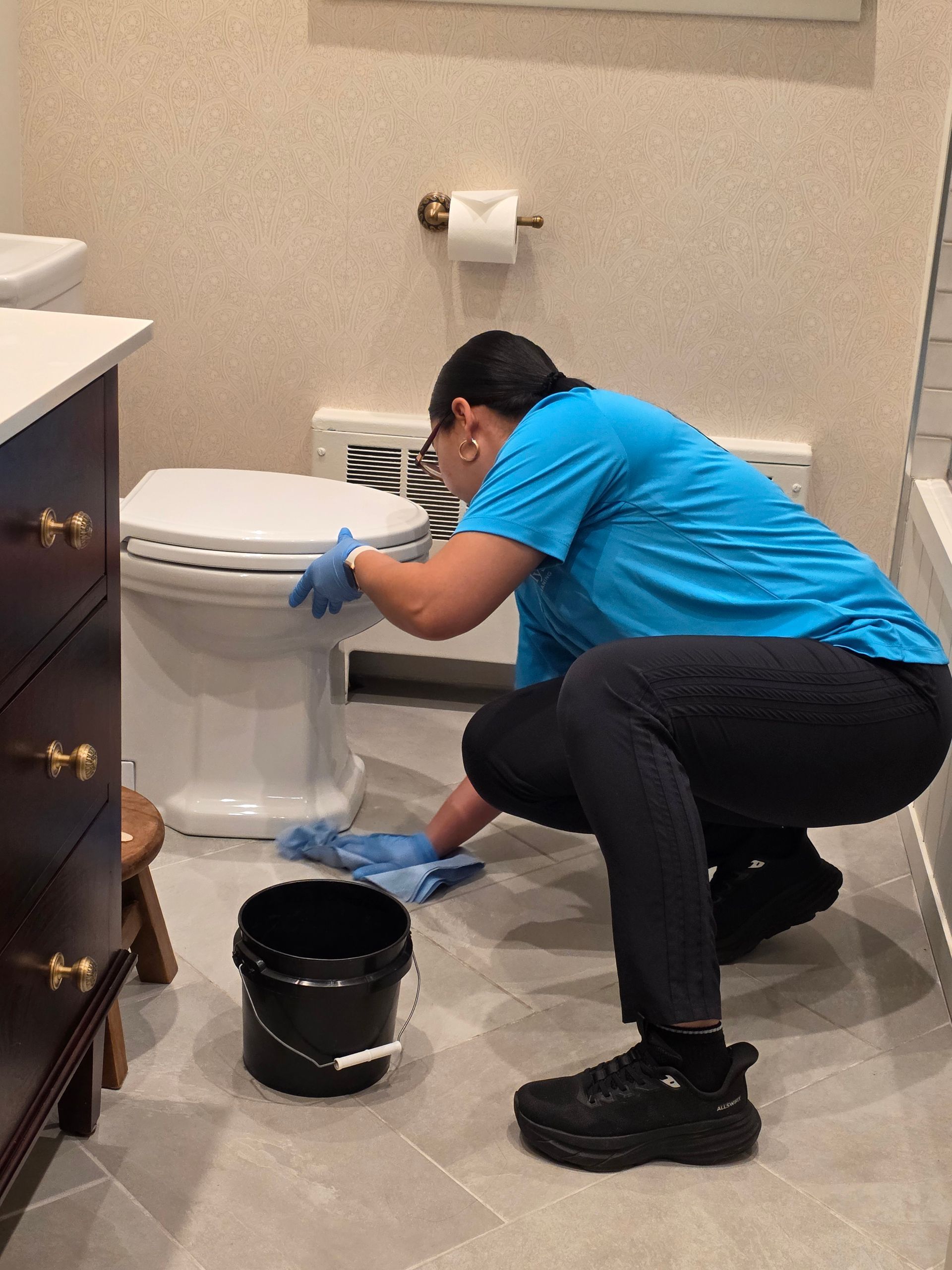Person in blue shirt cleaning a toilet in a bathroom, with a black bucket on the floor.