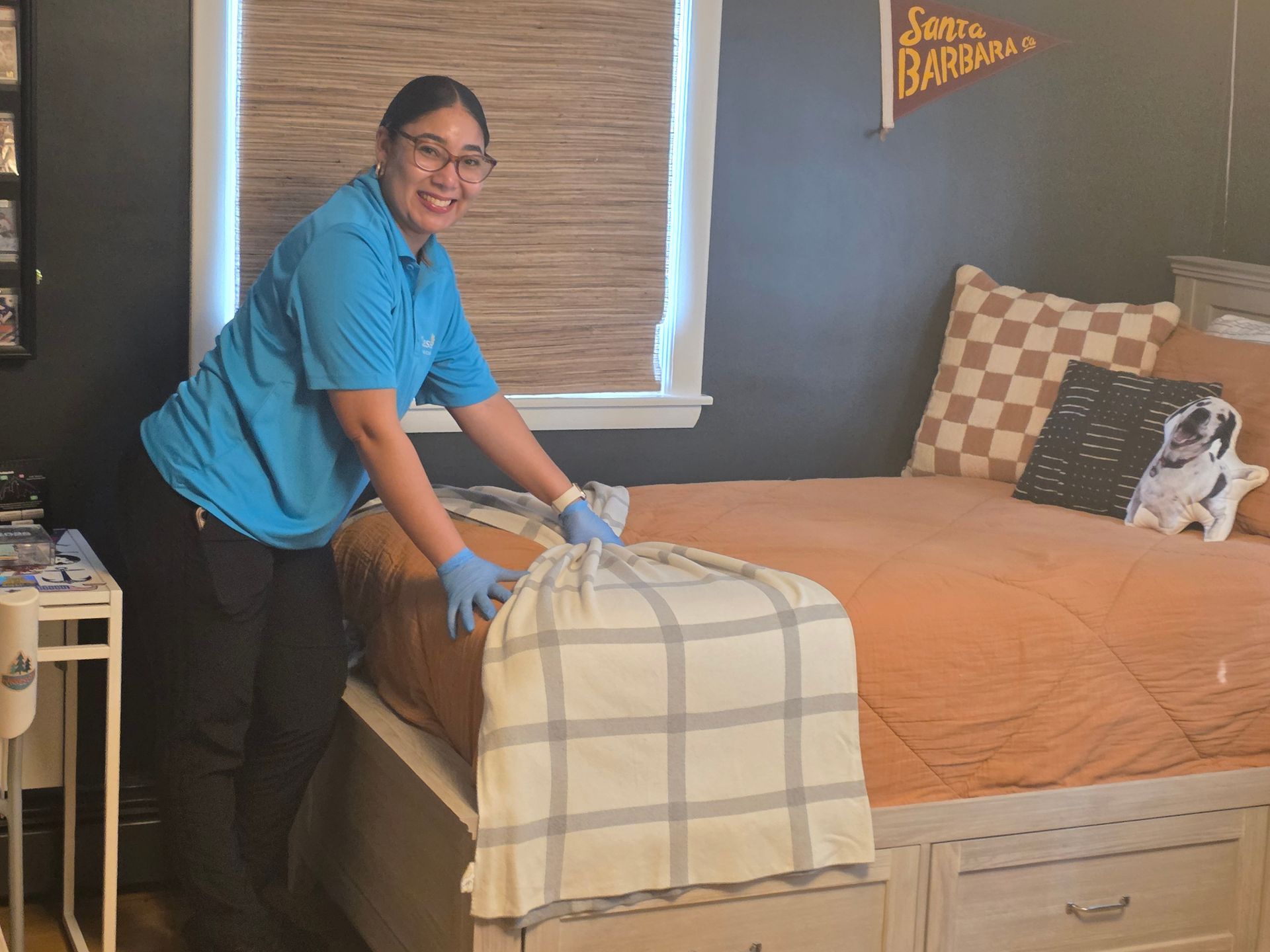 Person smiling and leaning over a bed in a bedroom with orange bedding and a plaid blanket.