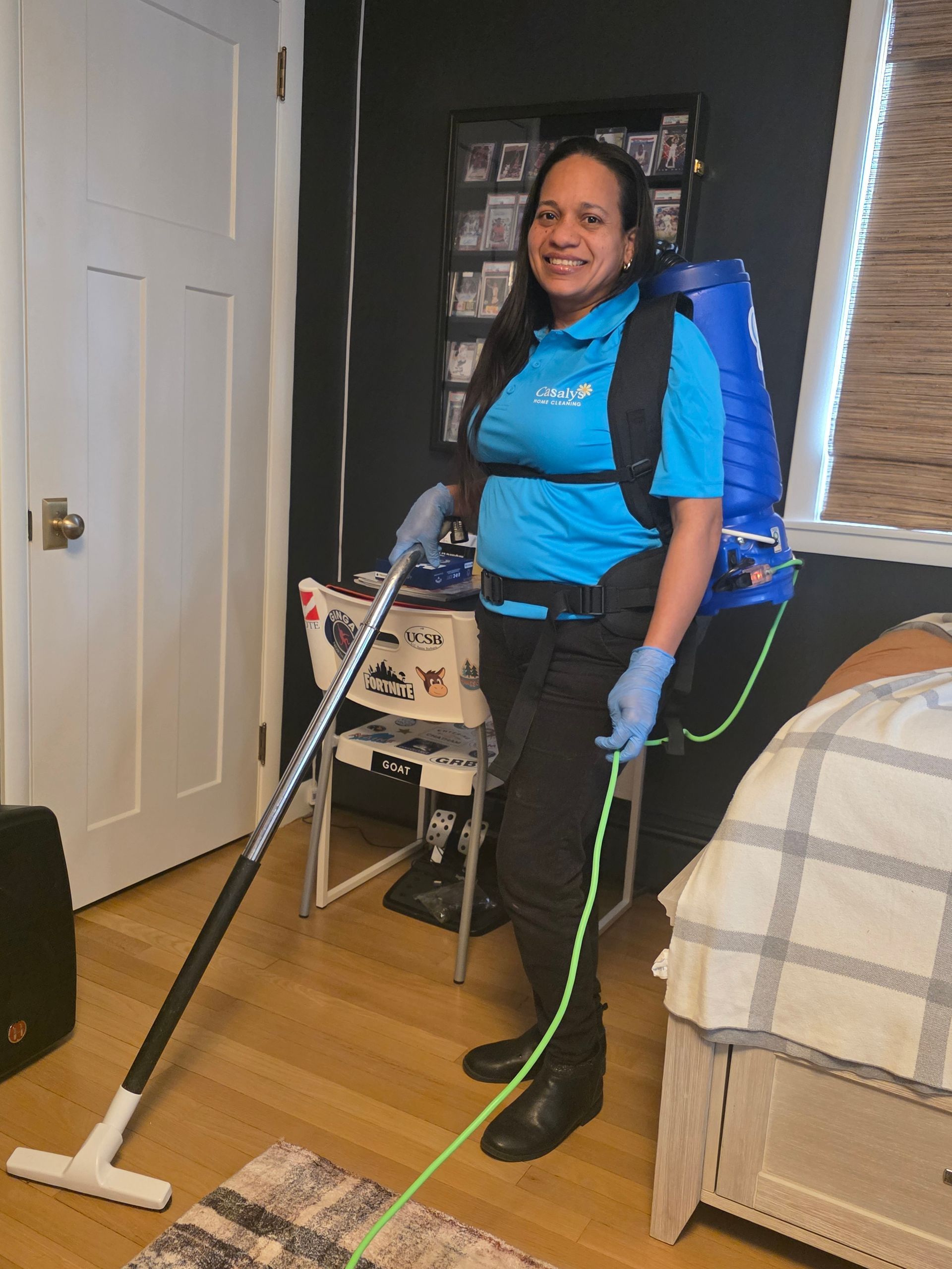 Person vacuuming a bedroom with a backpack vacuum and white floor nozzle, smiling near a bed and closet.