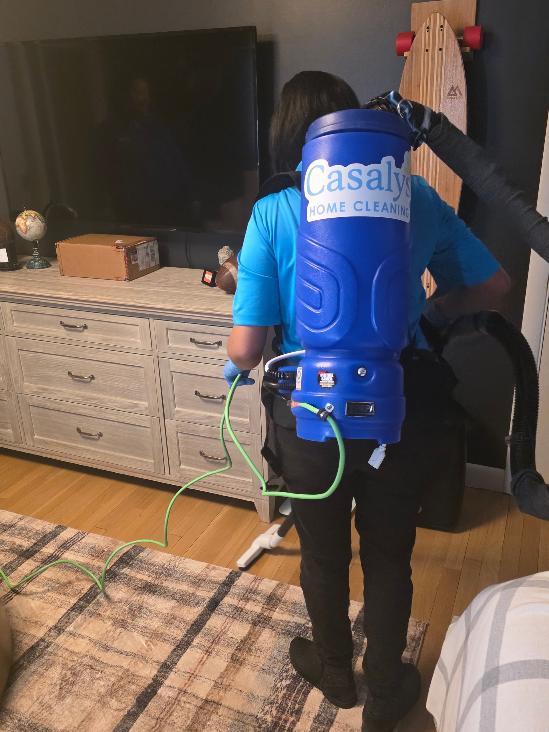 Person wearing a blue backpack sprayer in a living room, spraying near a TV stand.