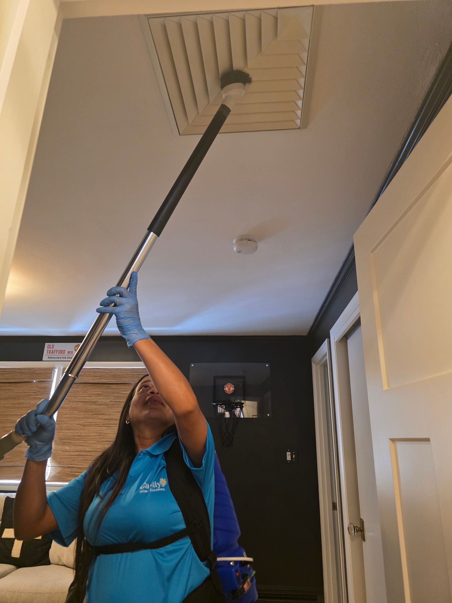 Person in blue gloves uses a long pole to clean a ceiling vent indoors.