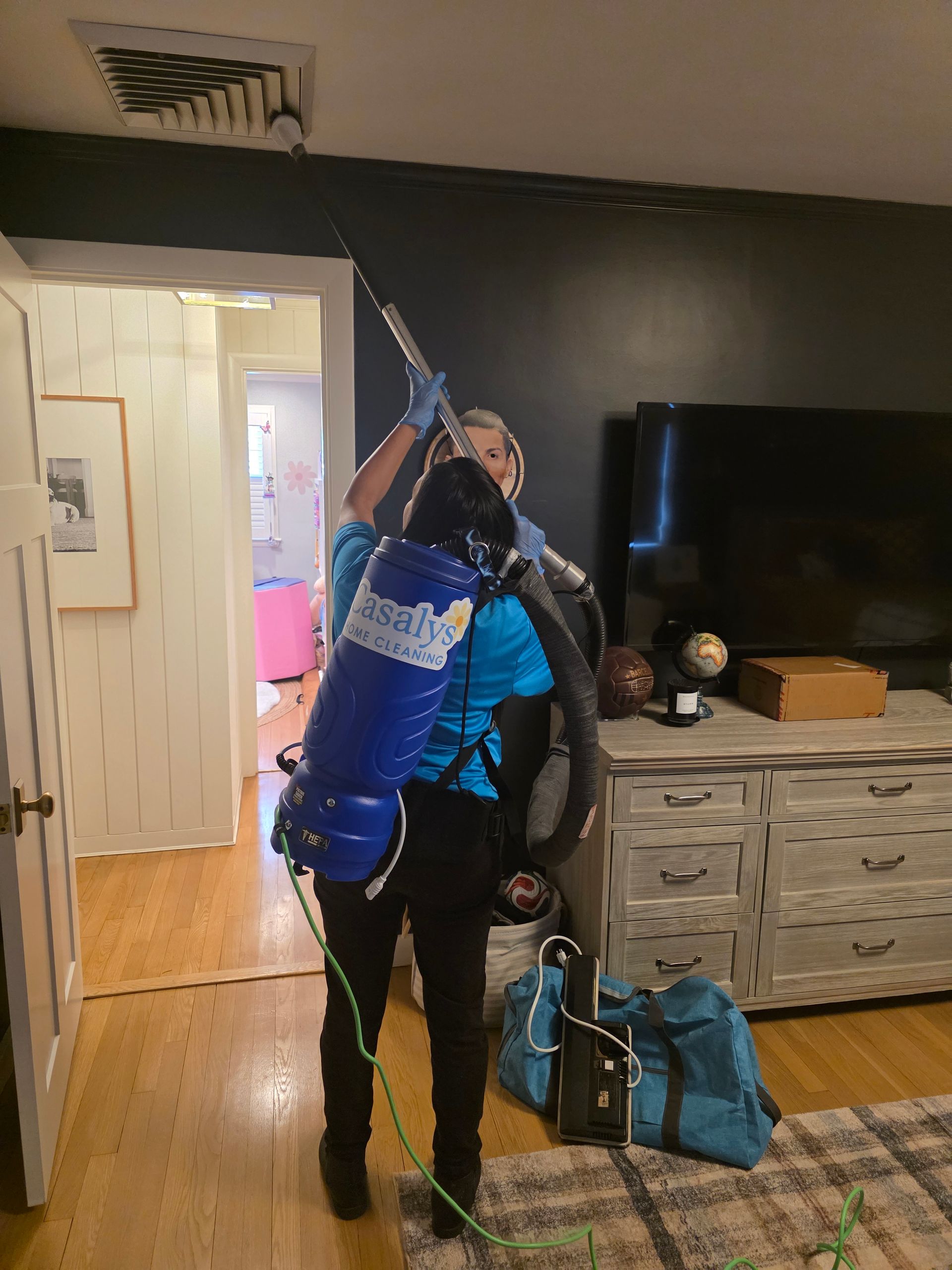 Person in a blue tank harnessing gear indoors, holding a long pole or tool, beside a TV and luggage.