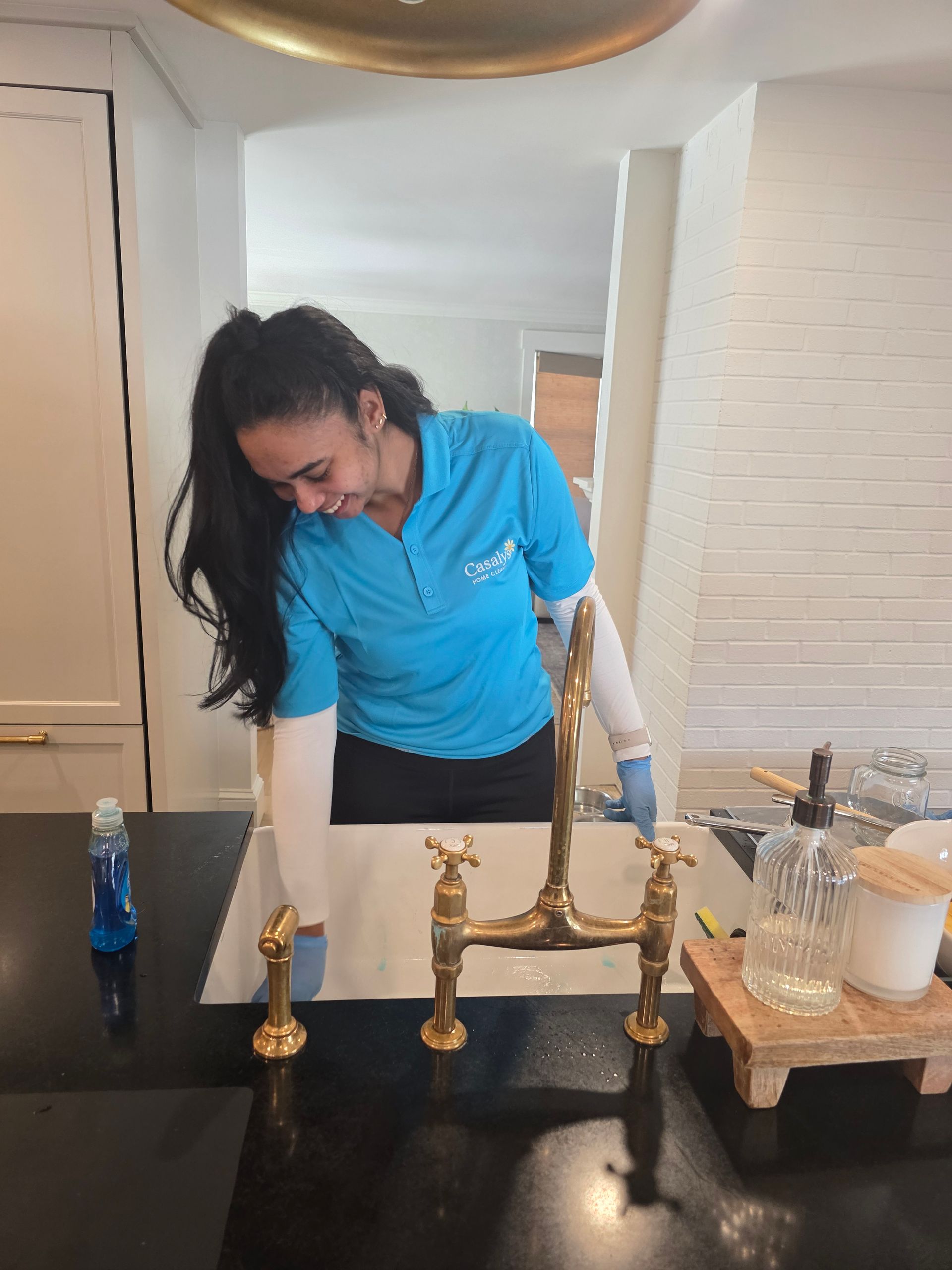 Person cleaning a black kitchen sink countertop with a spray bottle and cloth, beside a brass faucet