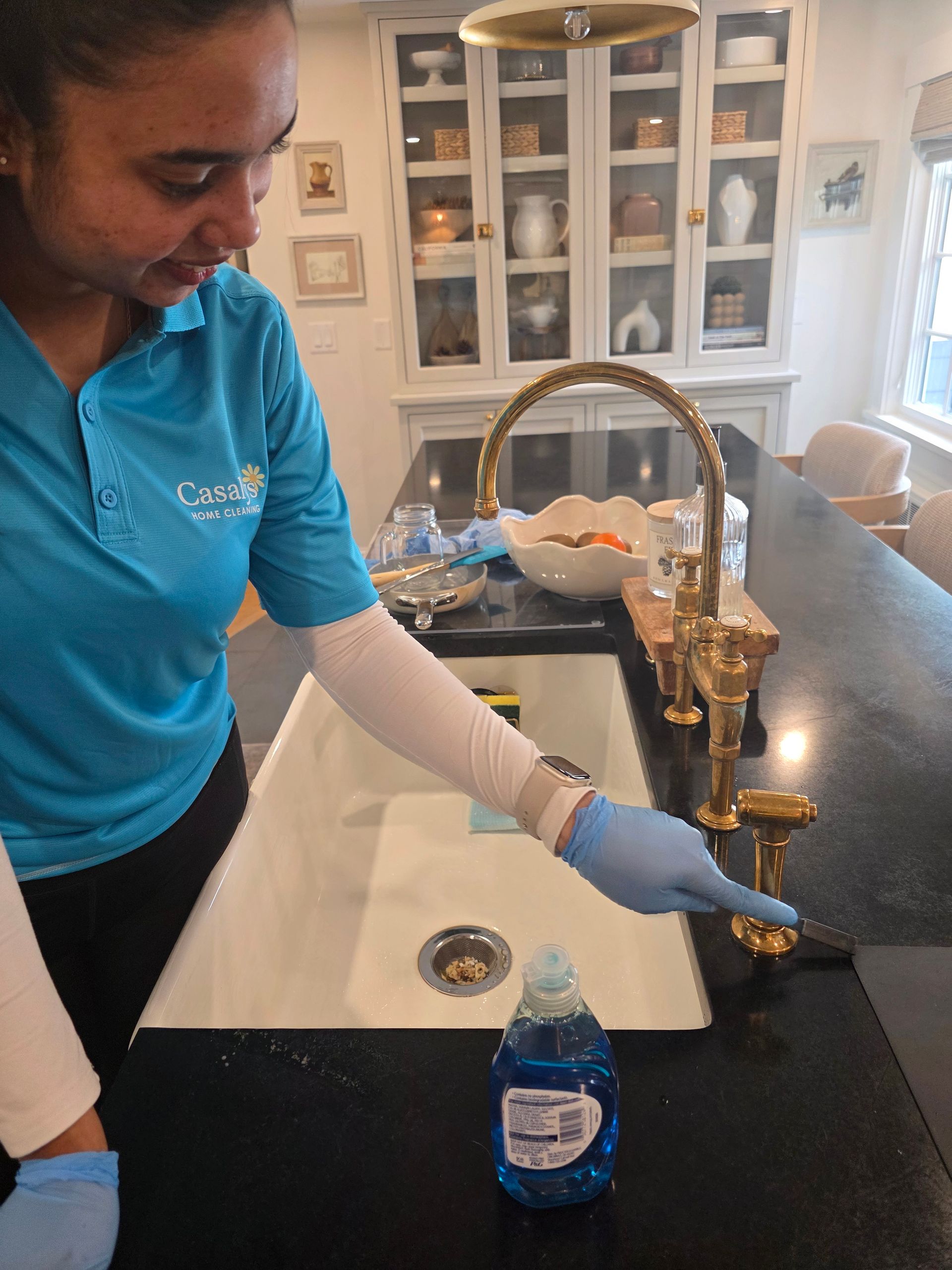 Person in blue gloves sprays and wipes a kitchen countertop near a sink and blue bottle.