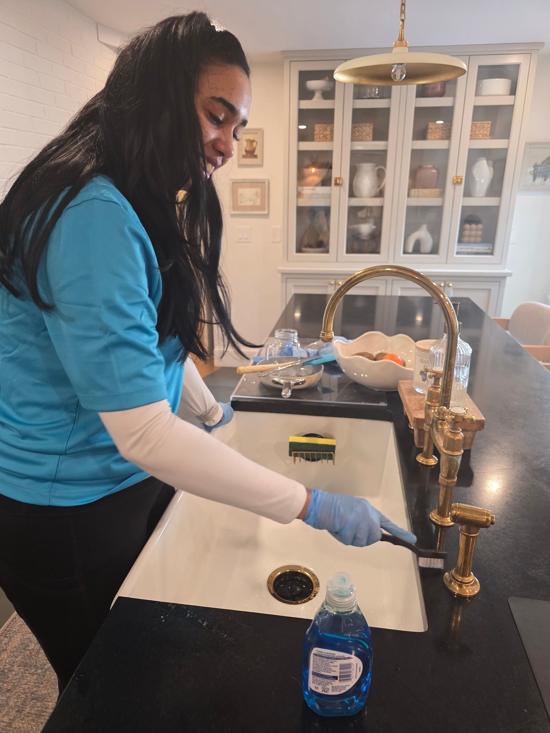 Woman in blue shirt and gloves cleaning a kitchen sink with a spray bottle and cloth.