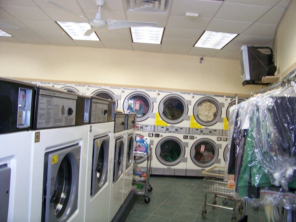A laundromat with multiple washers and dryers. Clothing hangs on a rack, lit by overhead fluorescent lights.